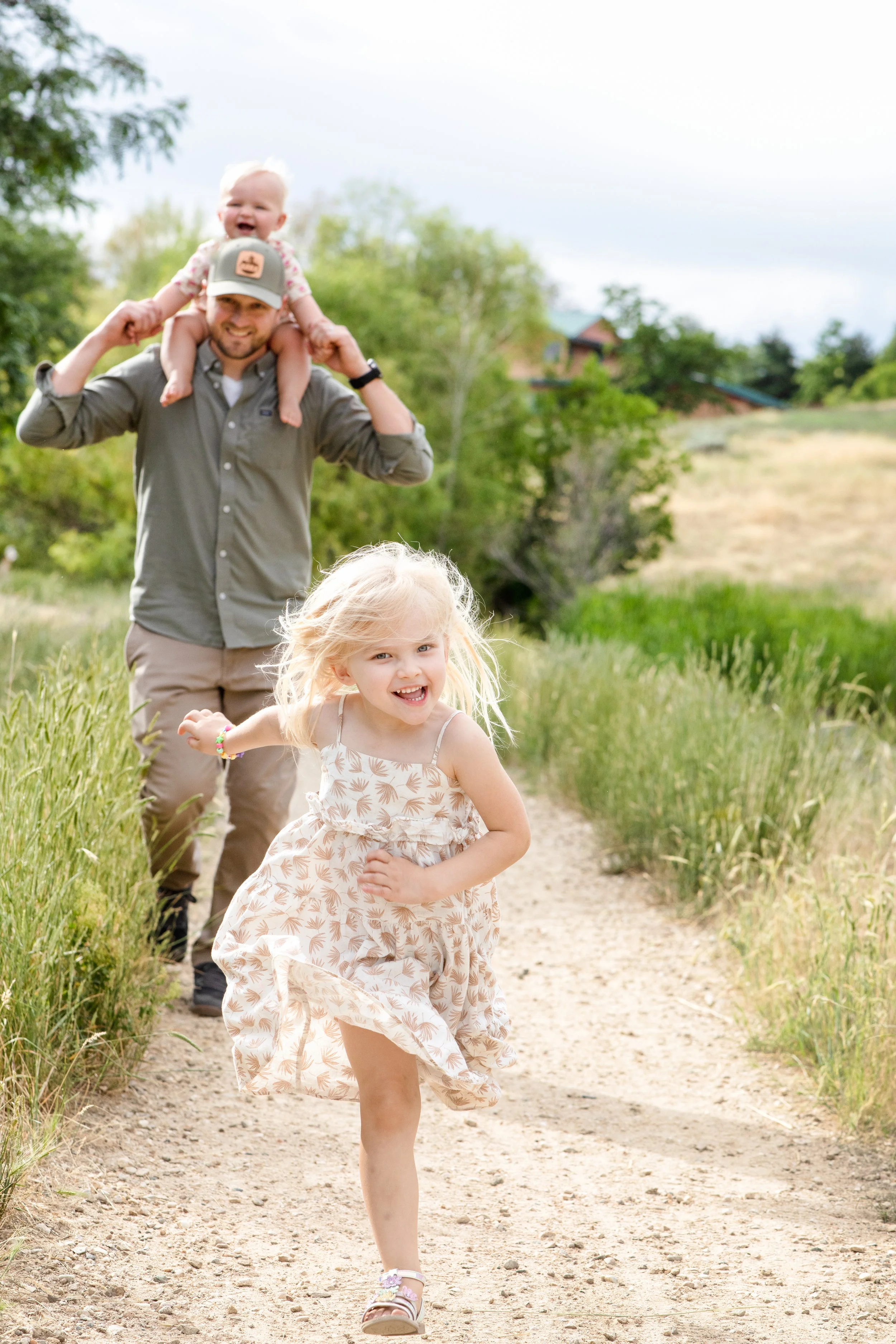 A family with a man, a little girl, and a baby, enjoying a walk outdoors on a dirt trail in a green, rural area with trees and grass, under partly cloudy skies.