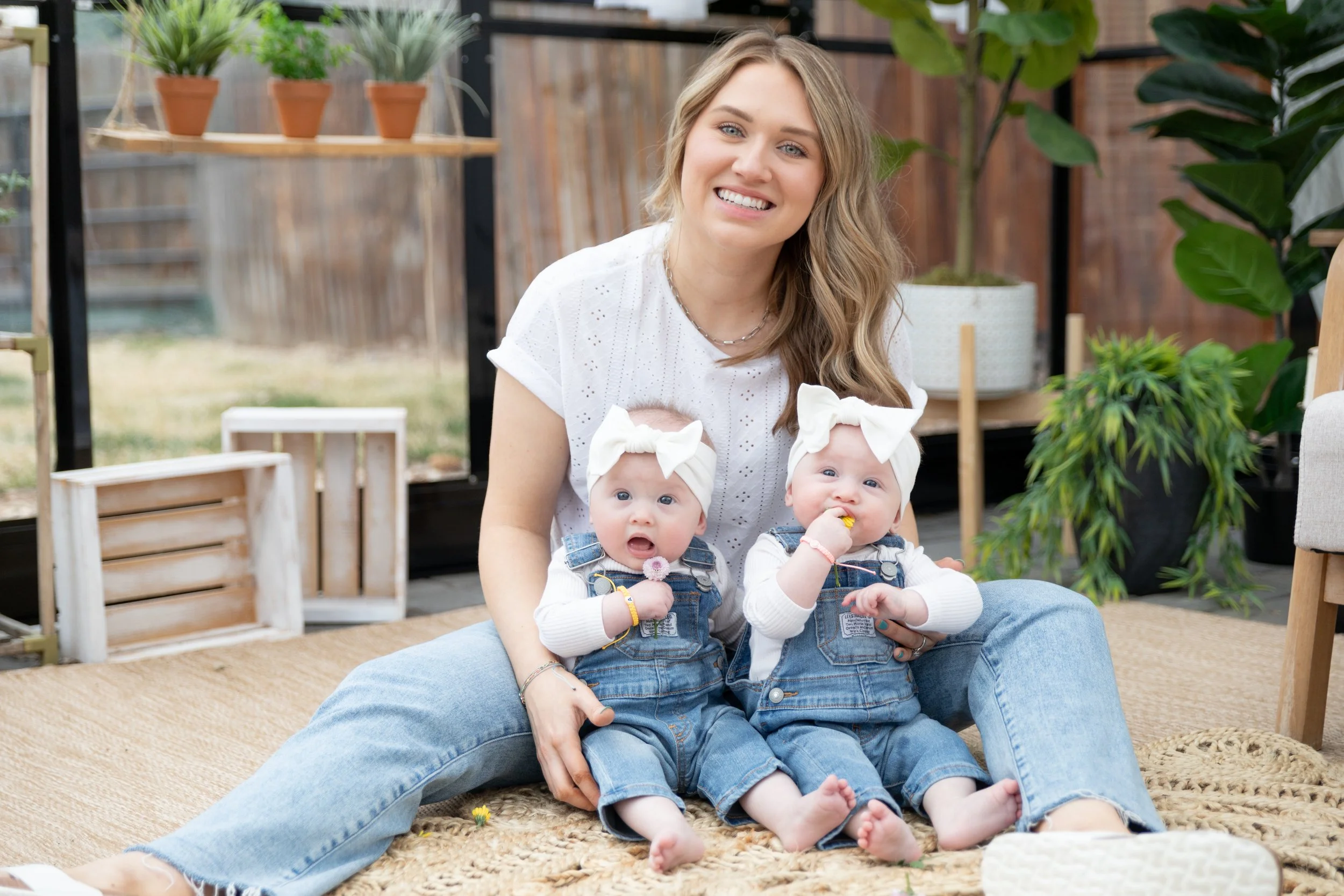 A woman sitting on the floor with two babies, all wearing jeans and white headbands with bows. The woman is smiling, and the babies are looking curious and one is eating a snack. The setting appears to be indoors with plants and shelves in the backgr