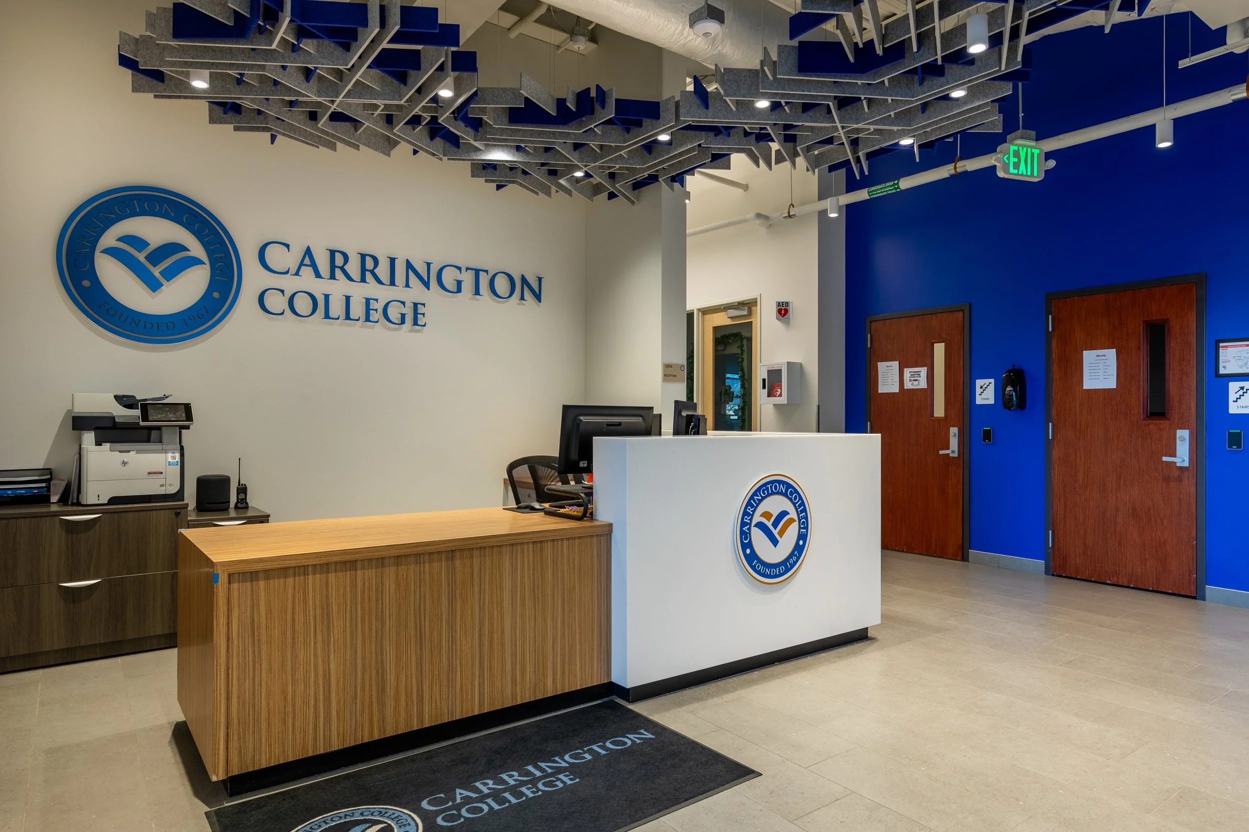 College reception area with a white front desk displaying the Carrington College logo, a wooden counter with office equipment, a light-colored wall with blue logo, blue and white walls with wooden doors, and ceiling acoustic panels.