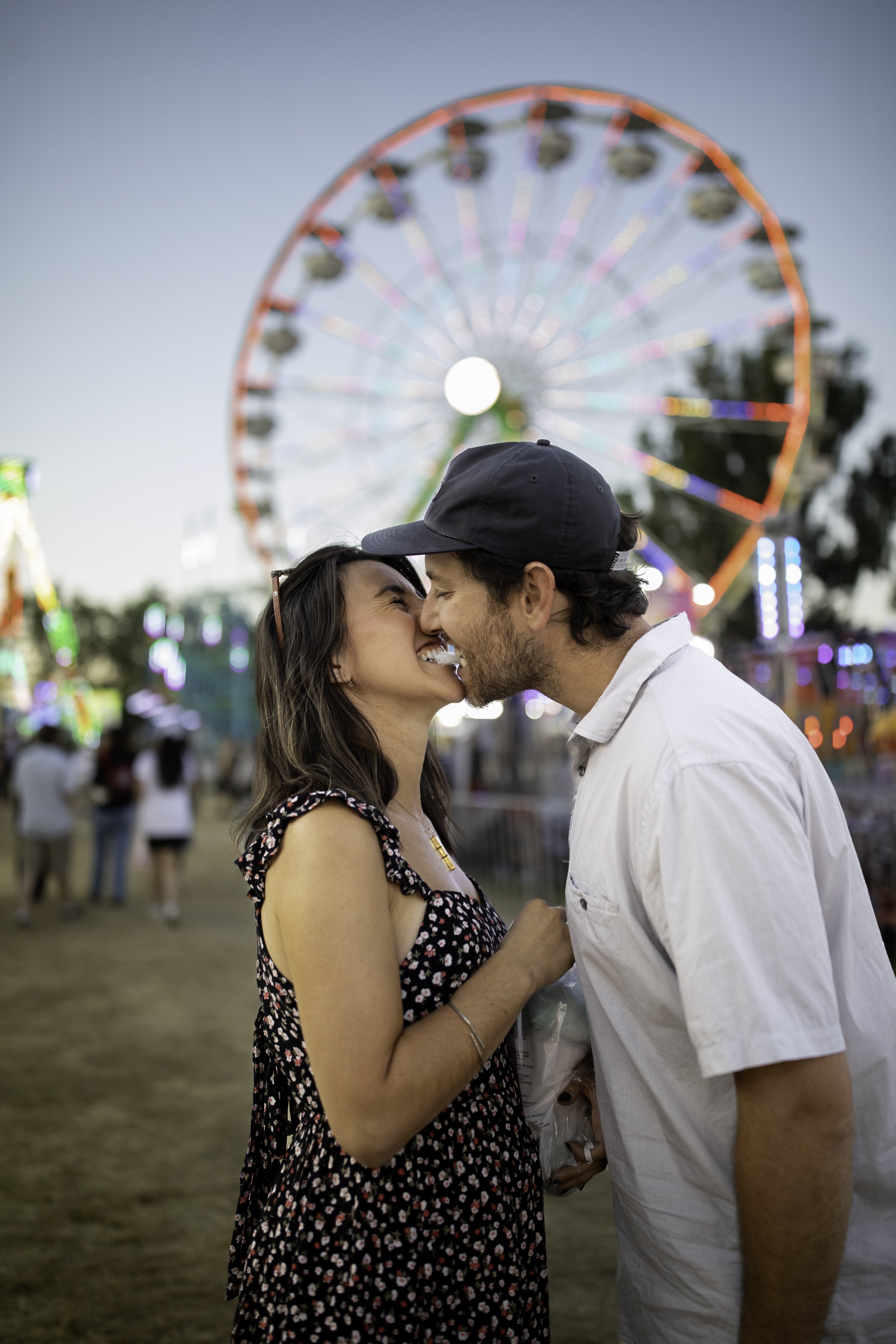 A couple sharing a kiss at a carnival during the evening with a Ferris wheel lit up in the background.