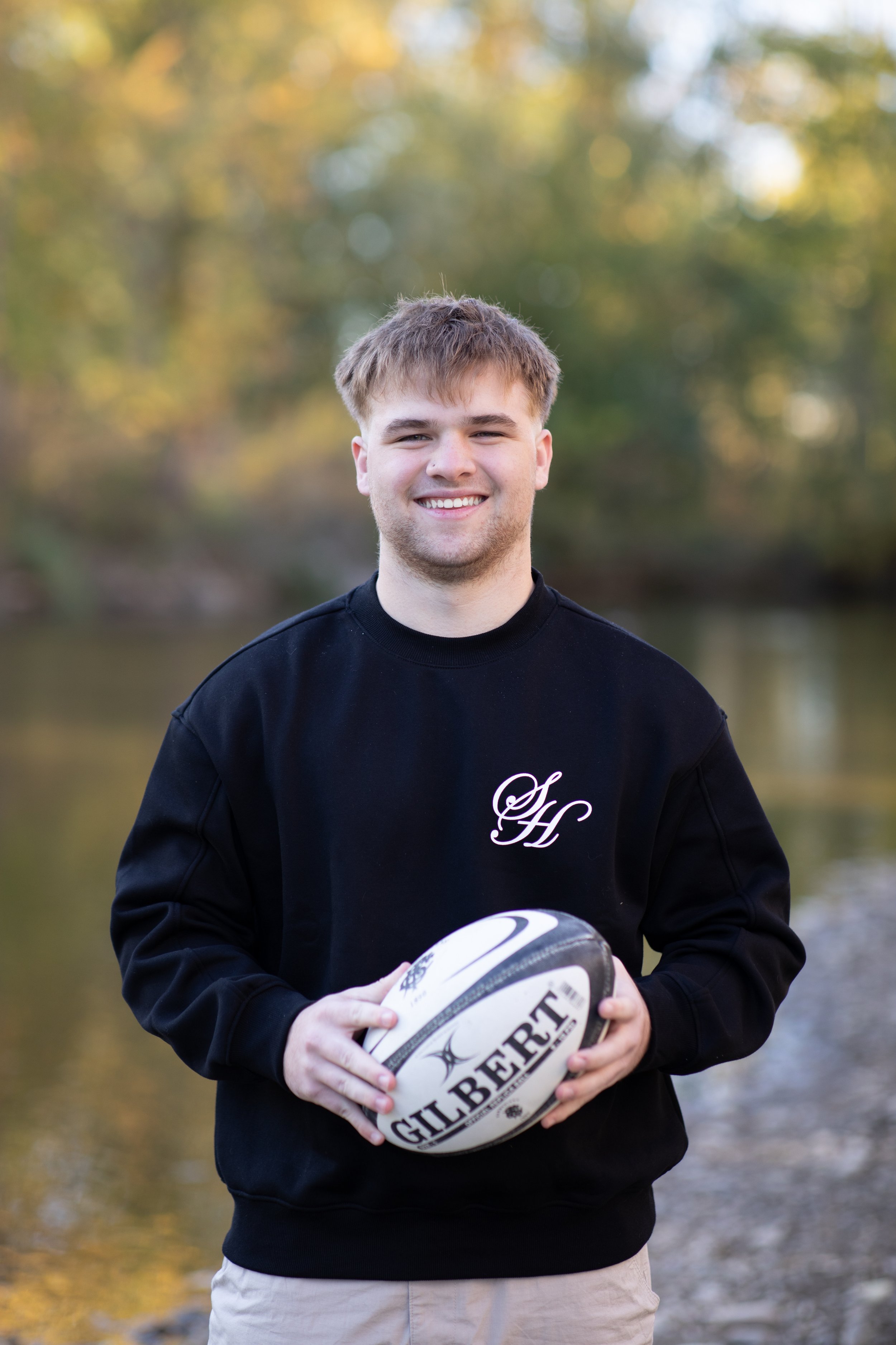 A young man with light brown hair, wearing a black sweatshirt with the initials 'SH' on it, is smiling and holding a rugby ball, standing outdoors near a body of water with trees in the background.