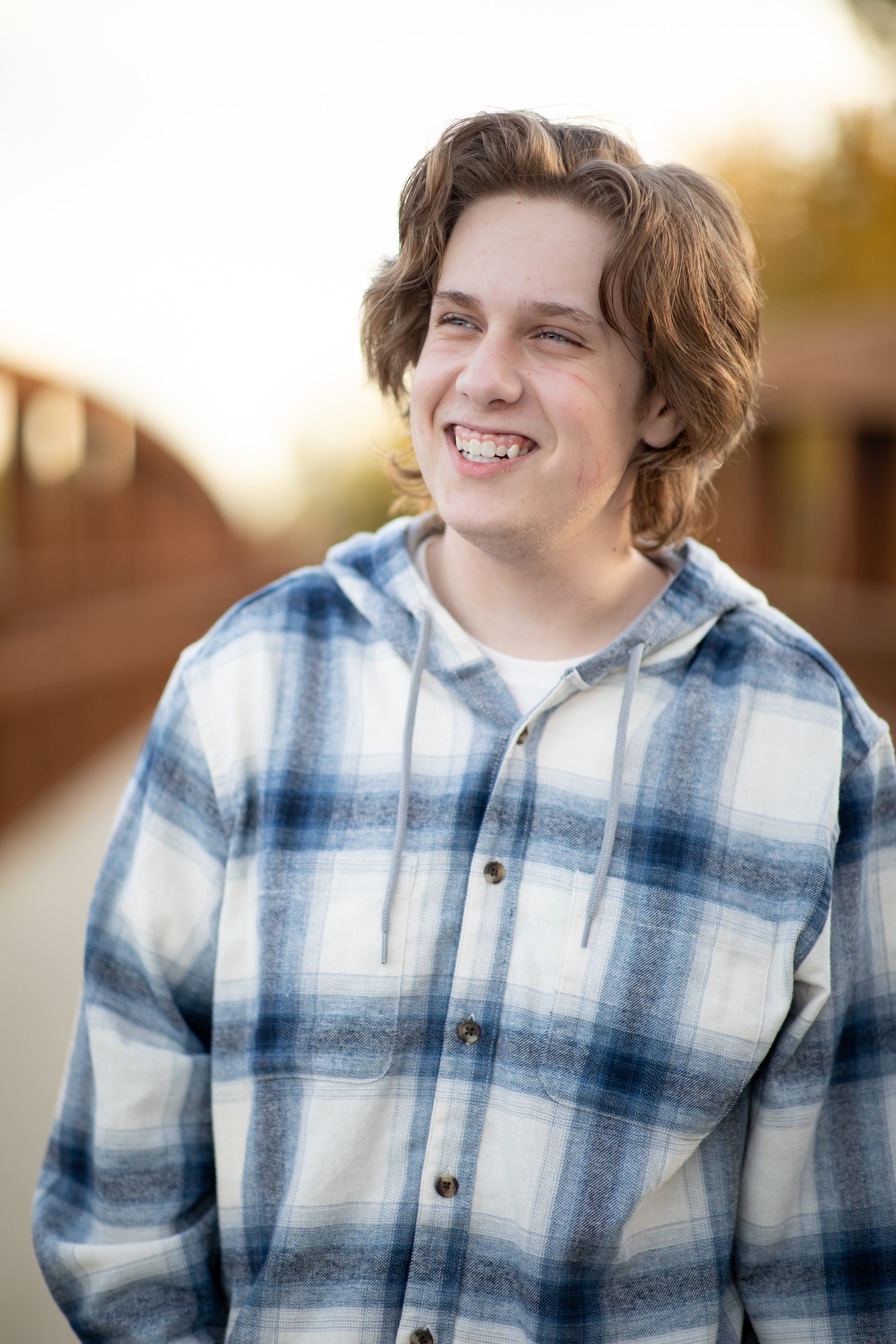 A young man with curly, medium-length hair smiling outdoors, wearing a blue and white plaid hoodie, with a wooden bridge and autumn foliage blurred in the background.