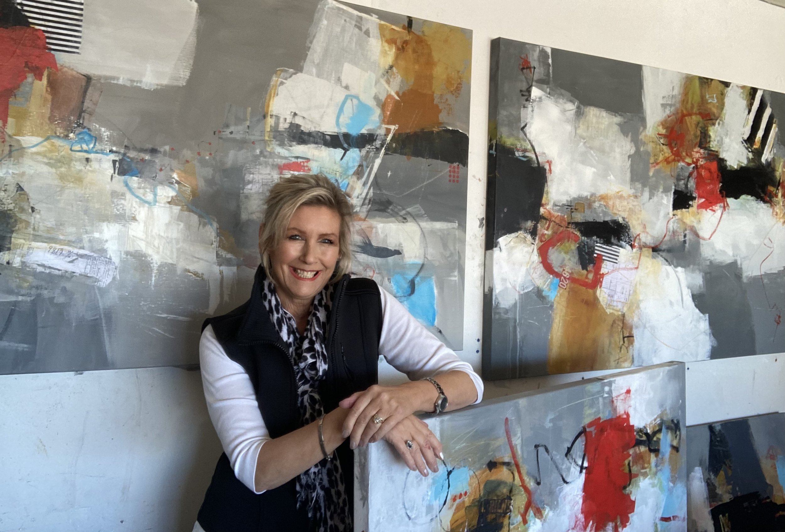 A woman with blonde hair and a leopard print scarf smiling, leaning on a surface with multiple abstract paintings in shades of gray, black, white, red, blue, and gold behind her.