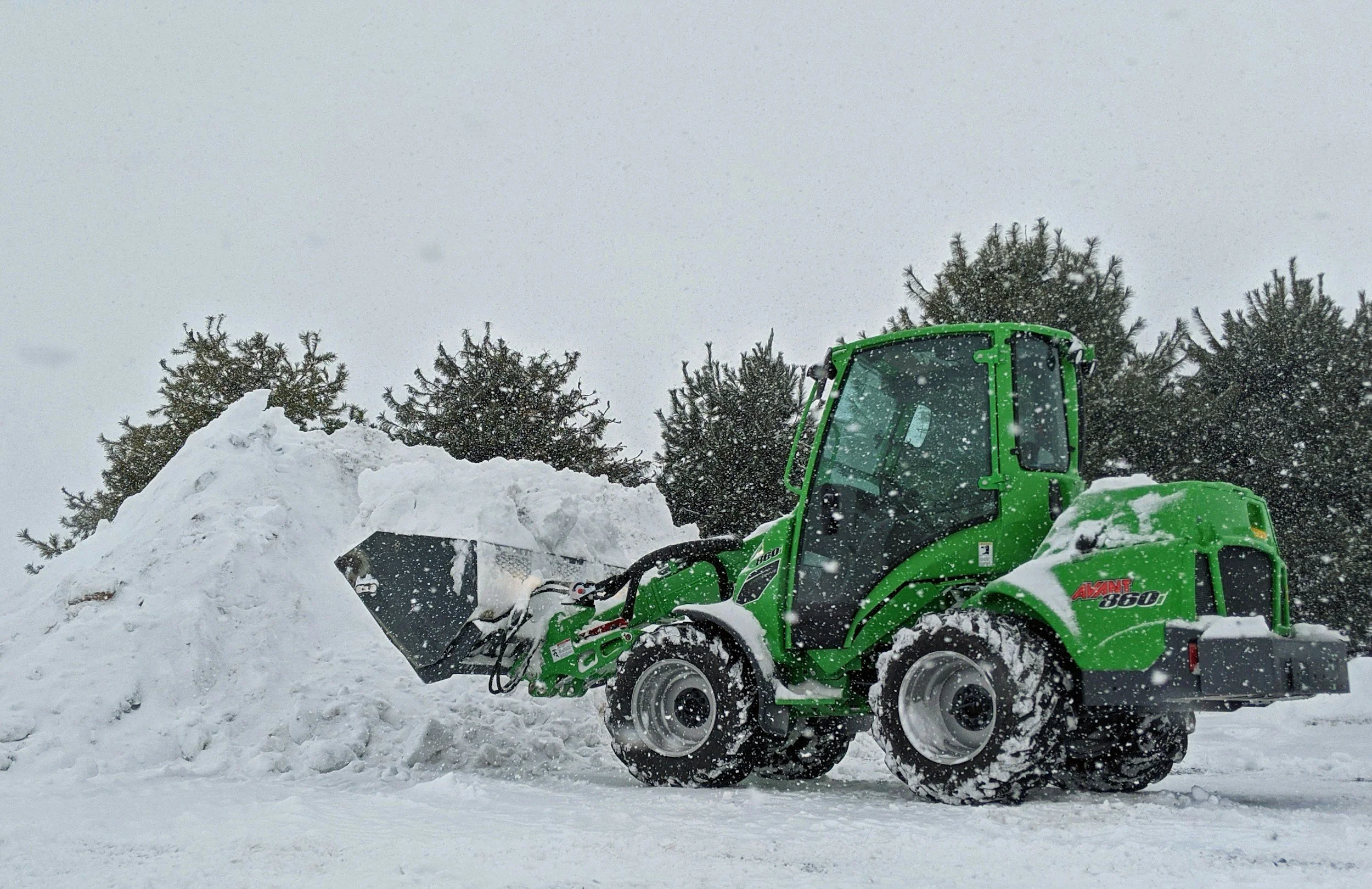 Green compact skid steer loader clearing snow in a snowy outdoor area with trees in the background.