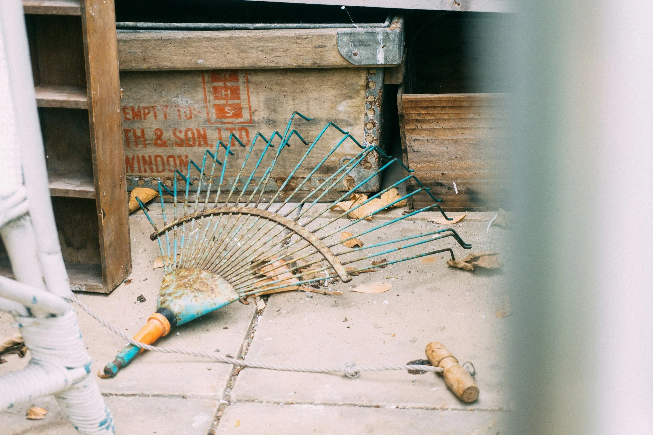 A rusted garden rake with a broken handle, lying on the concrete floor among fallen leaves, in front of wooden shelves and a wooden crate in a cluttered space.