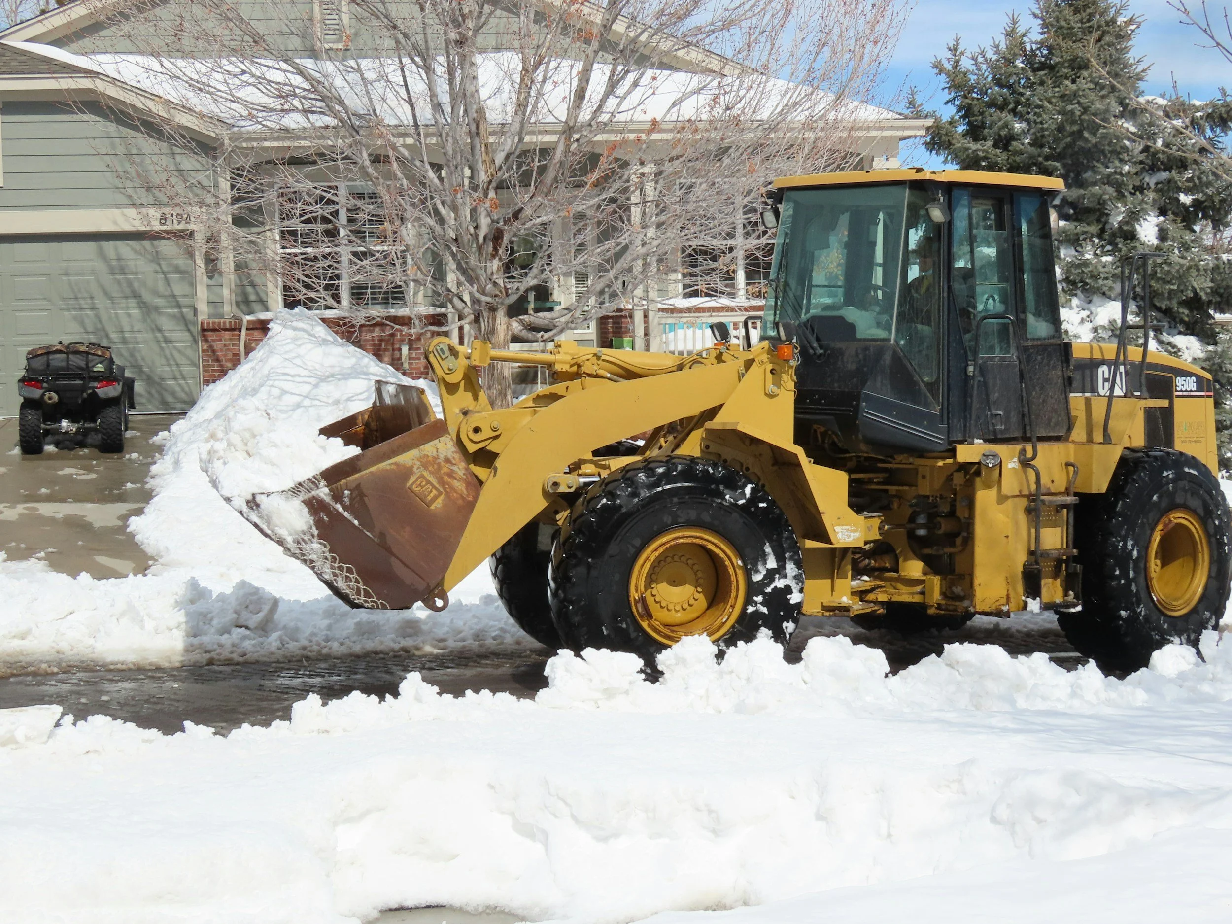 A yellow Caterpillar backhoe loader clearing snow from a driveway in a residential neighborhood. Snow is piled on the side and trees are leafless, indicating winter.