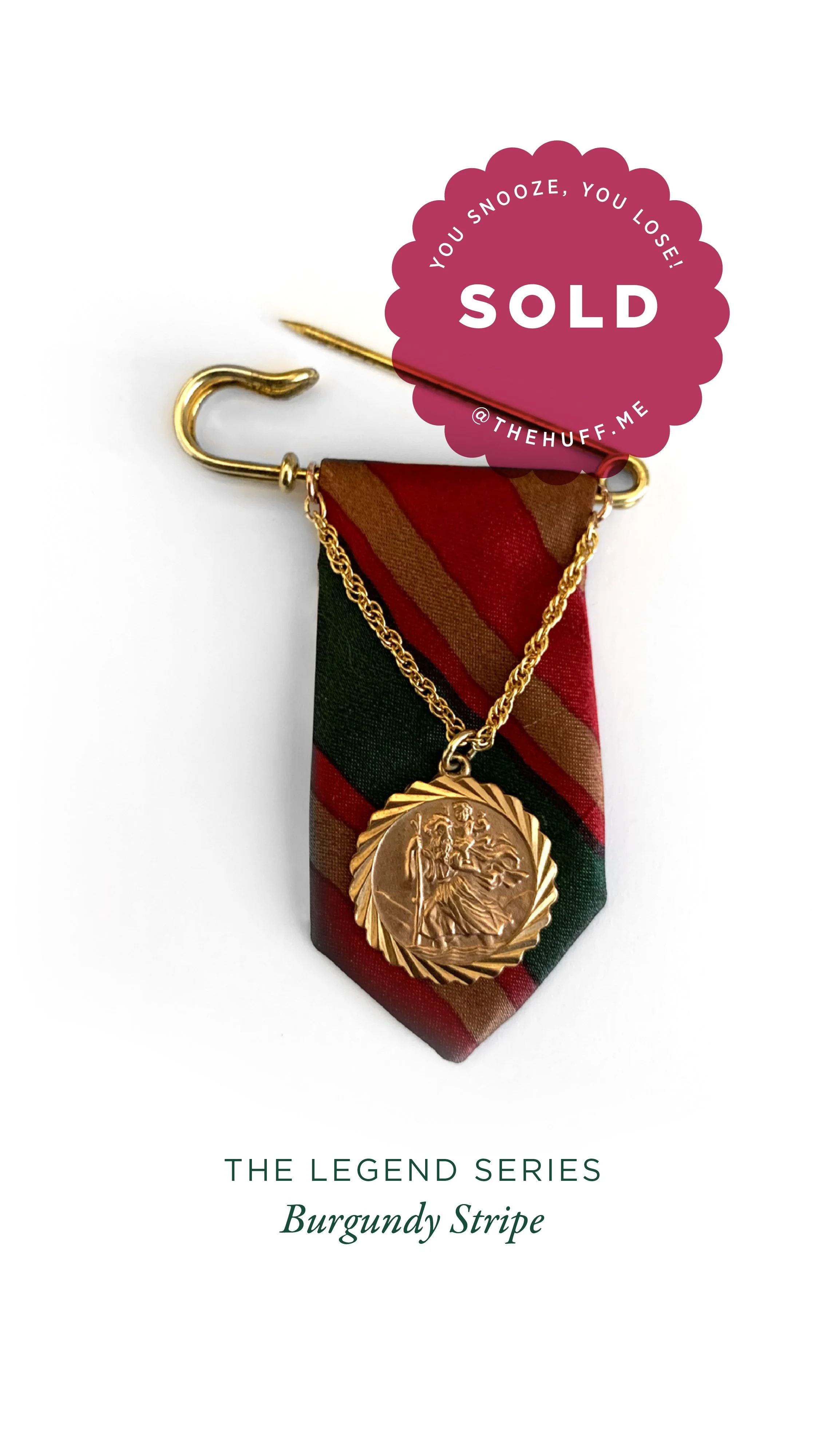 A gold brooch with a circular medallion featuring a detailed engraved figure, attached to a burgundy and green striped fabric tie, with a gold chain connecting the brooch and the tie, and a pink sticker that says 'SOLD.'