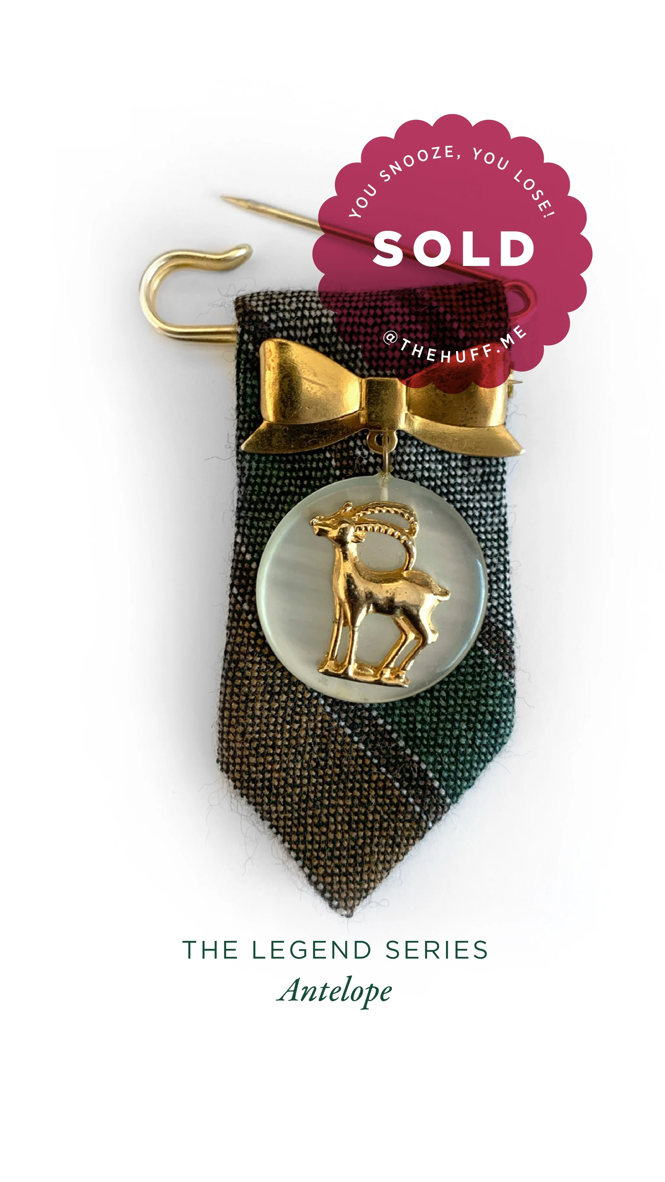 A tie pin with a gold antelope charm, attached to a plaid fabric background with a gold bow and safety pin, labeled as 'The Legend Series Antelope' and marked sold with a pink sticker that says 'You snooze, you lose!'