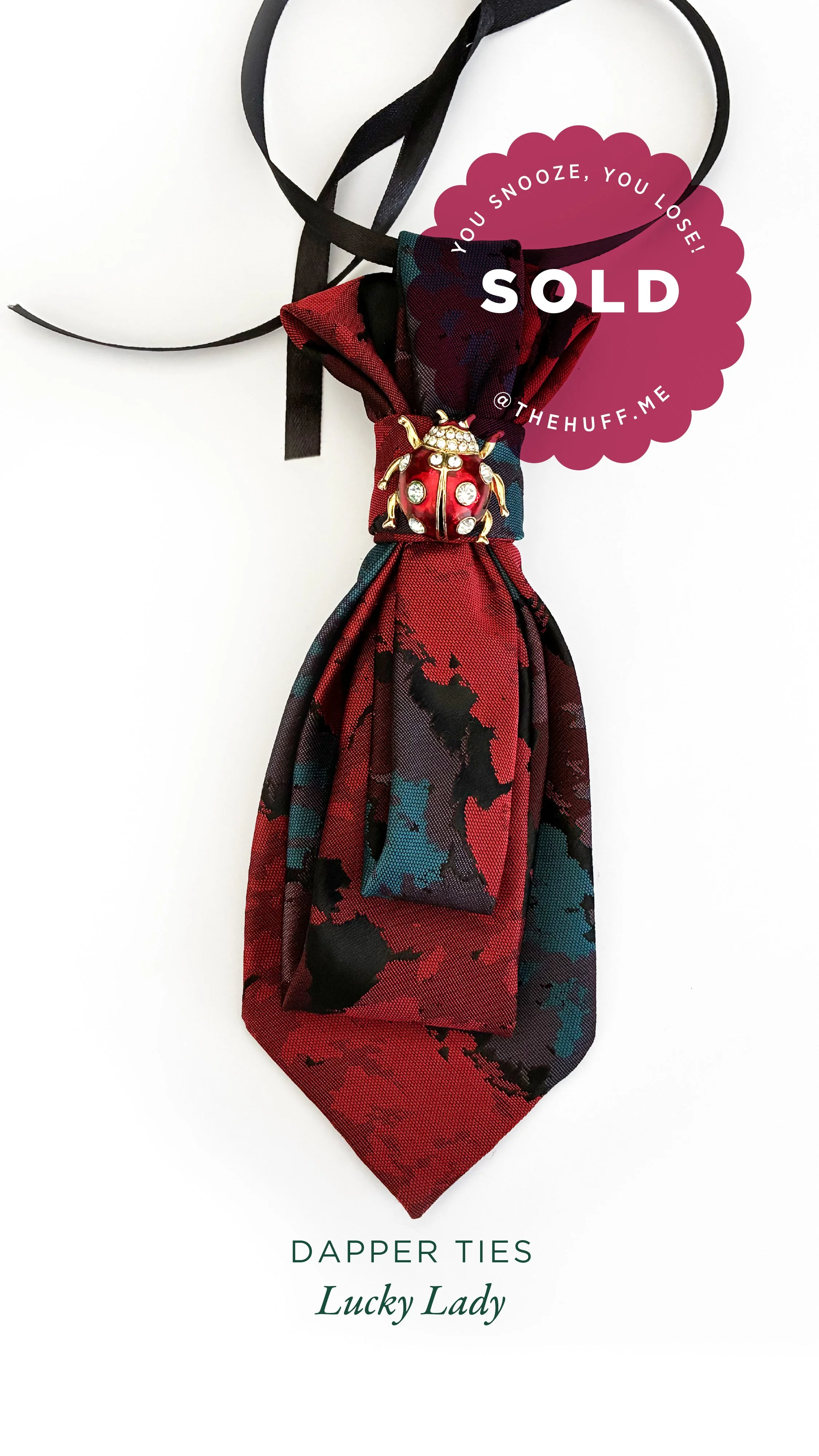 Red, black, and teal patterned silk dapper tie with a gold ladybug pin and a black ribbon, sold by The Huff.