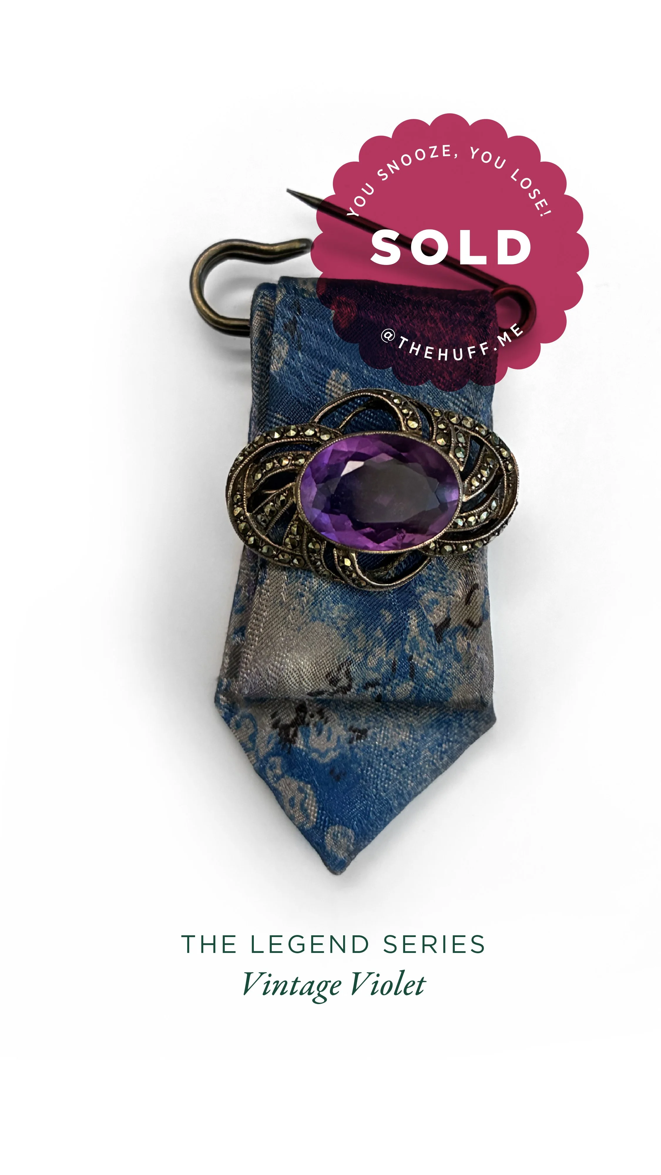 A jewellery brooch with a large purple gemstone, placed on a fabric tie blade with a decorative pattern. A pink sticker indicates the item is sold and warns against snoozing or losing it.