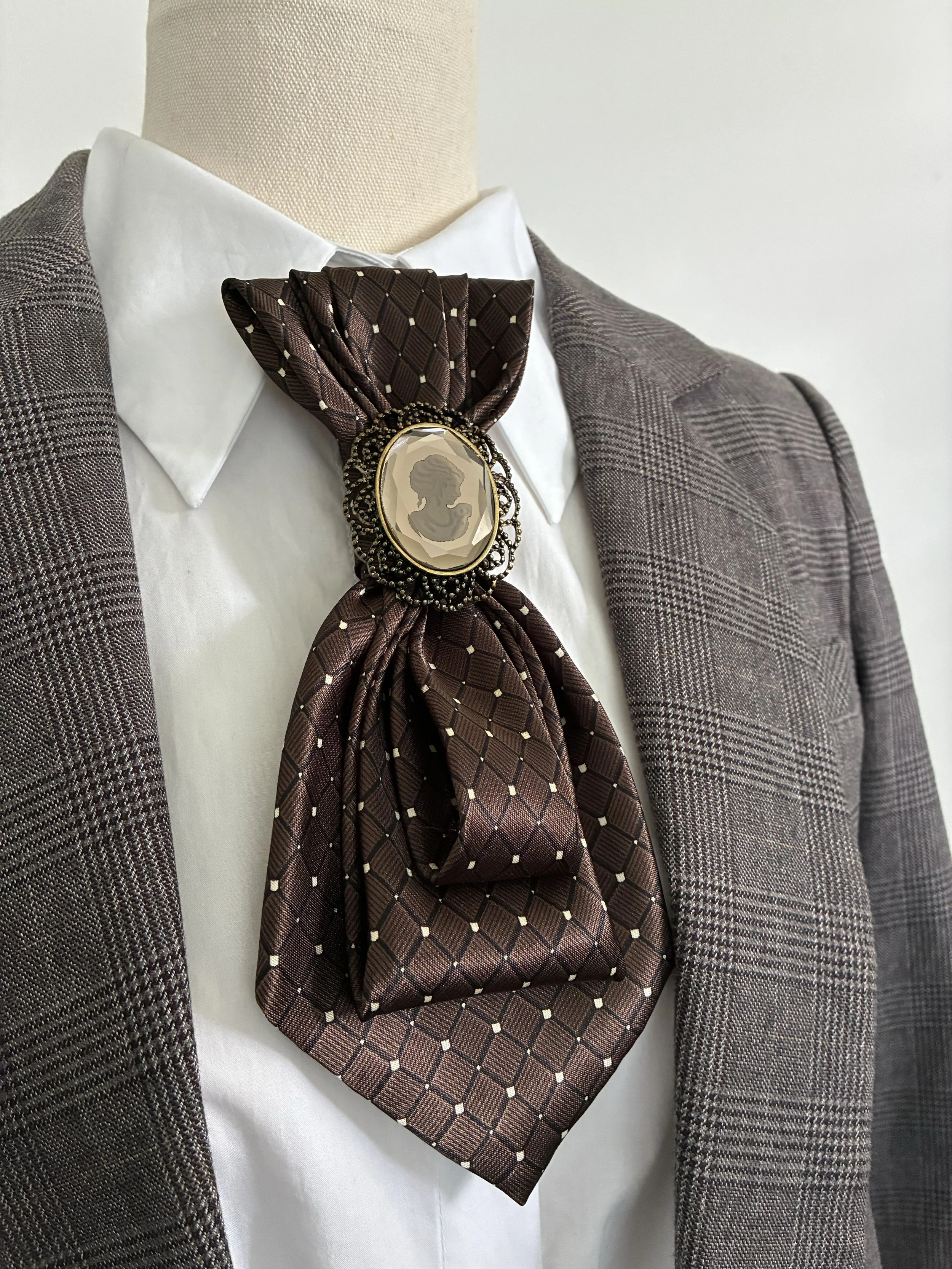 Close-up of a formal women's outfit on a mannequin, featuring a brown checkered blazer, white dress shirt, and a brown patterned silk jabot with a cameo brooch.