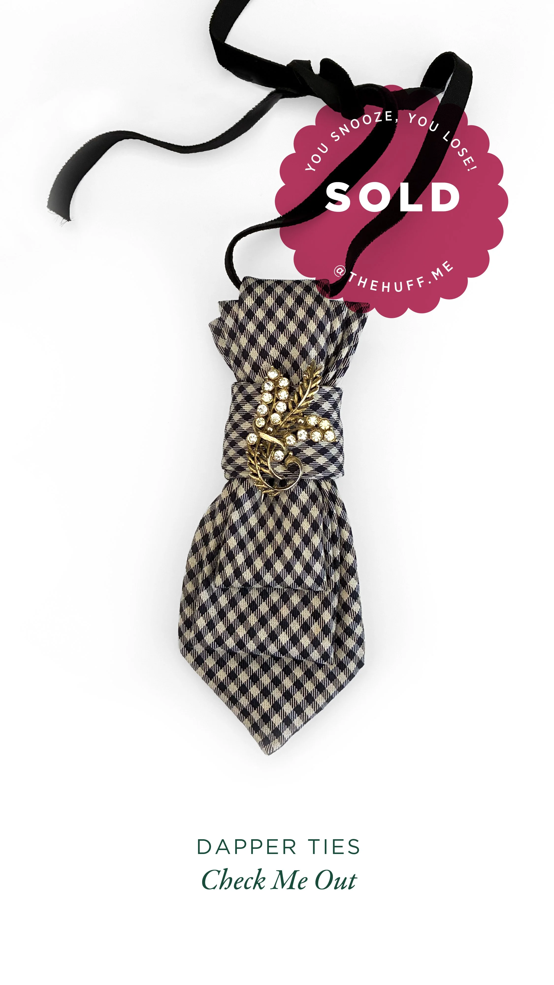 A black and white checkered dapper tie with a gold decorative brooch attached, displayed on a white background, with a pink sale sticker indicating the item is sold.