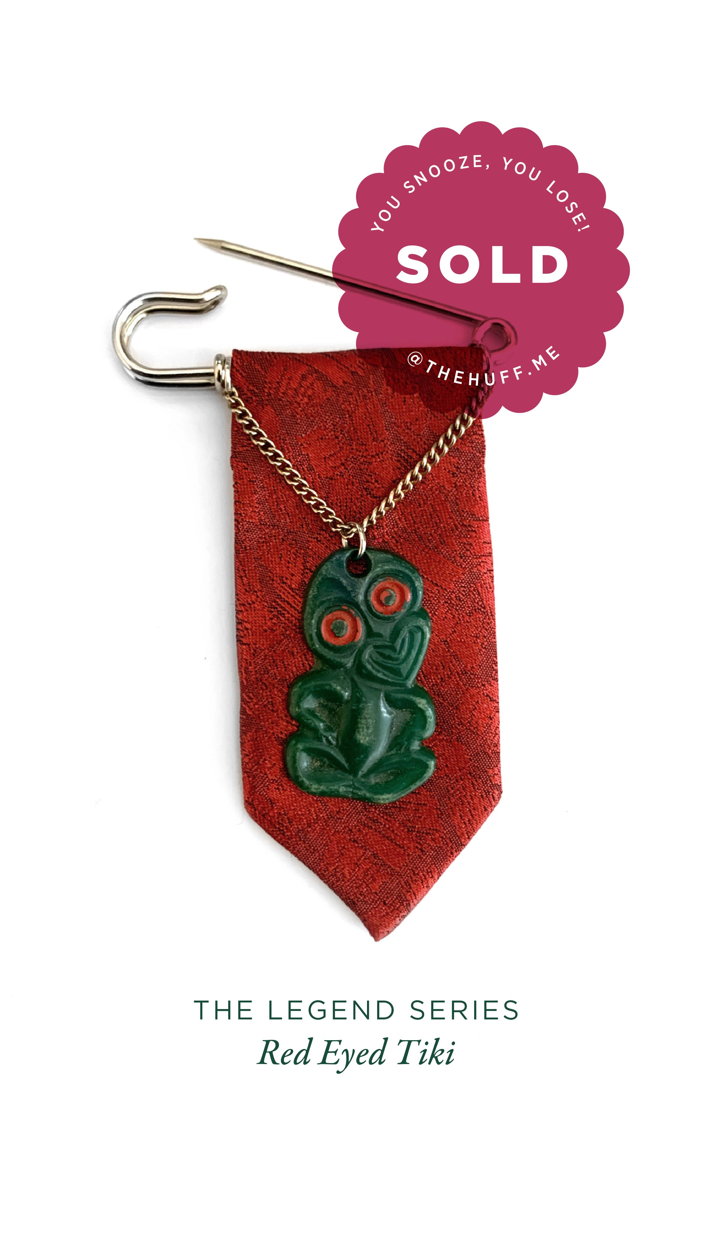 A green tiki necklace with red eyes on a red fabric backing, pinned with a safety pin, with a "Sold" tag indicating it is part of The Legend Series called Red Eyed Tiki.