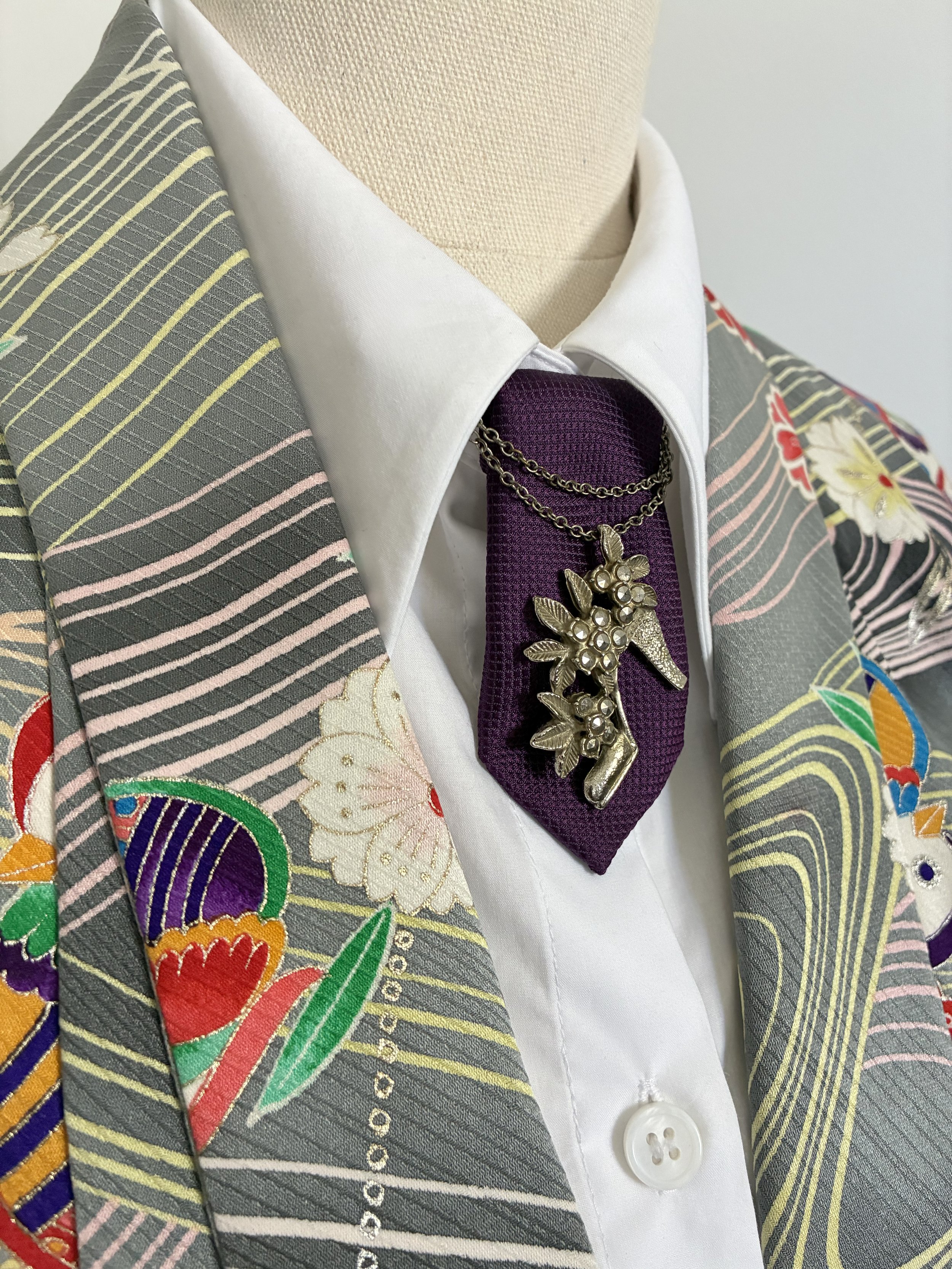 A mannequin dressed in a white shirt, a purple tie, a brooch with leaves, pearls, and a shoe, all layered over a kimono-style garment with colorful floral and stripe patterns.
