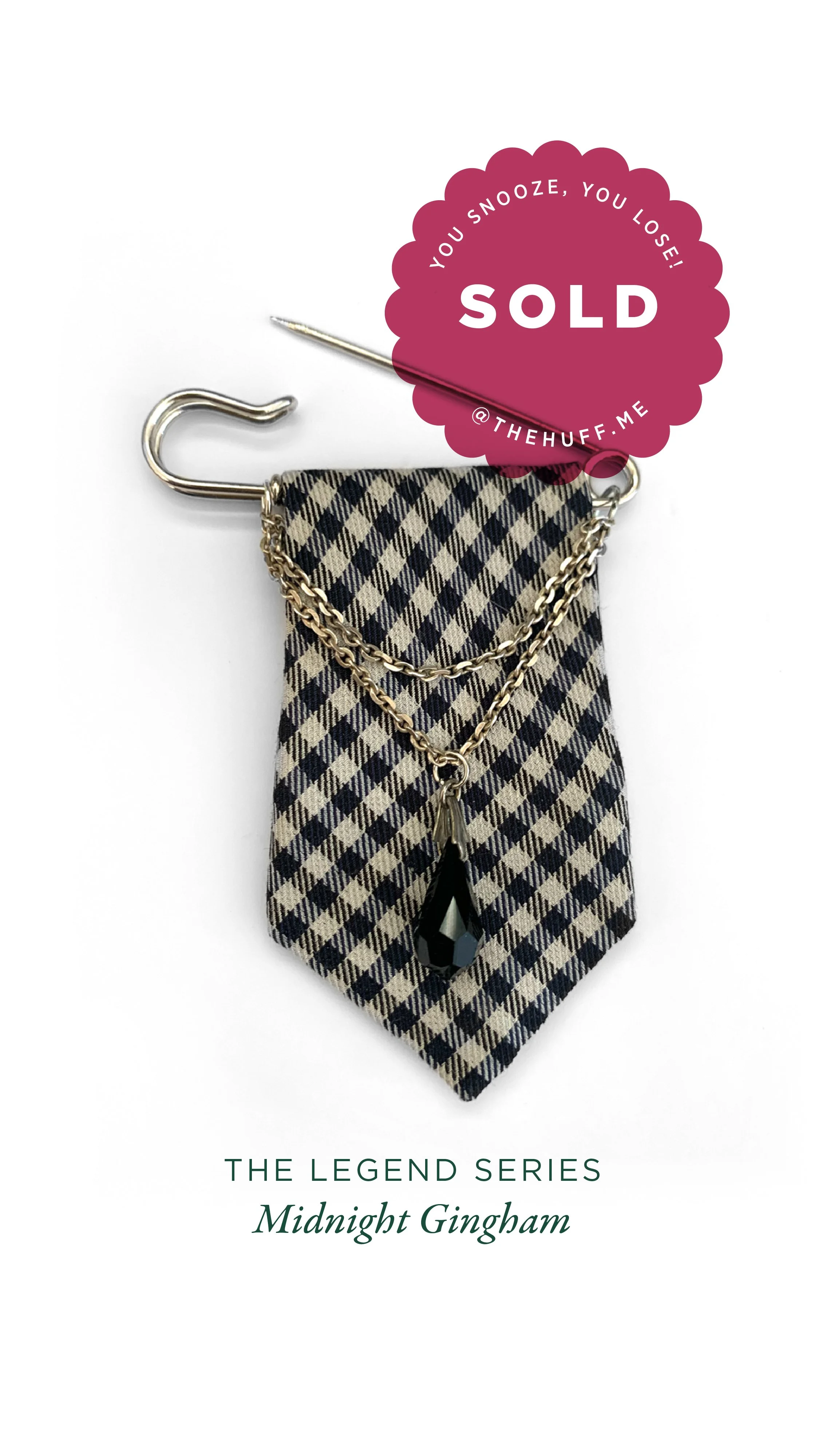 A jewellery pin featuring layered gold chains and a black crystal pendant, set against a checkered fabric tie blade with a 'SOLD' sticker on top.