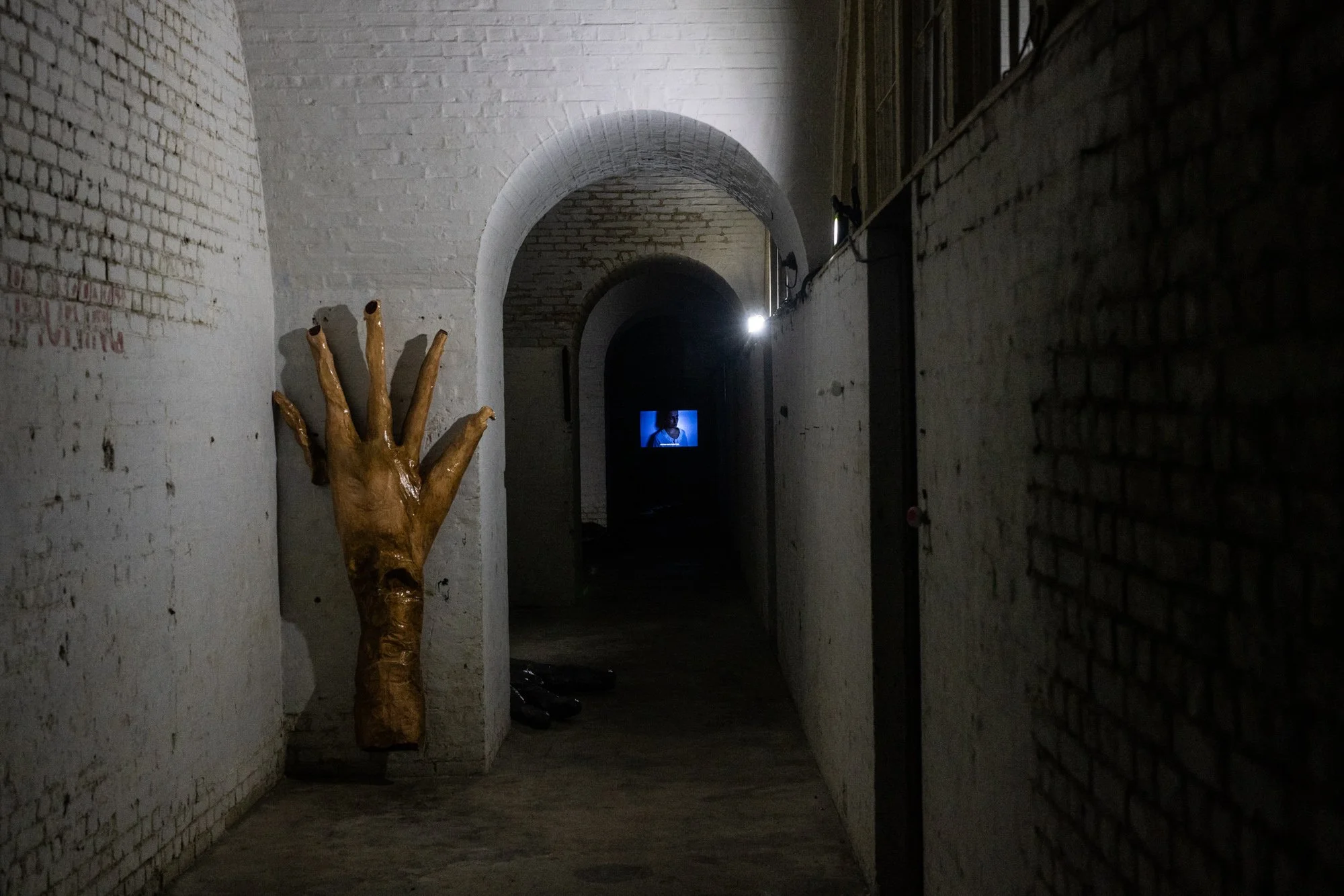 A dark hallway in an industrial or art gallery setting with brick walls and arched ceilings. There is a golden sculpture of a hand with elongated fingers on the left side of the hallway, and a small television screen displaying a person in the distan