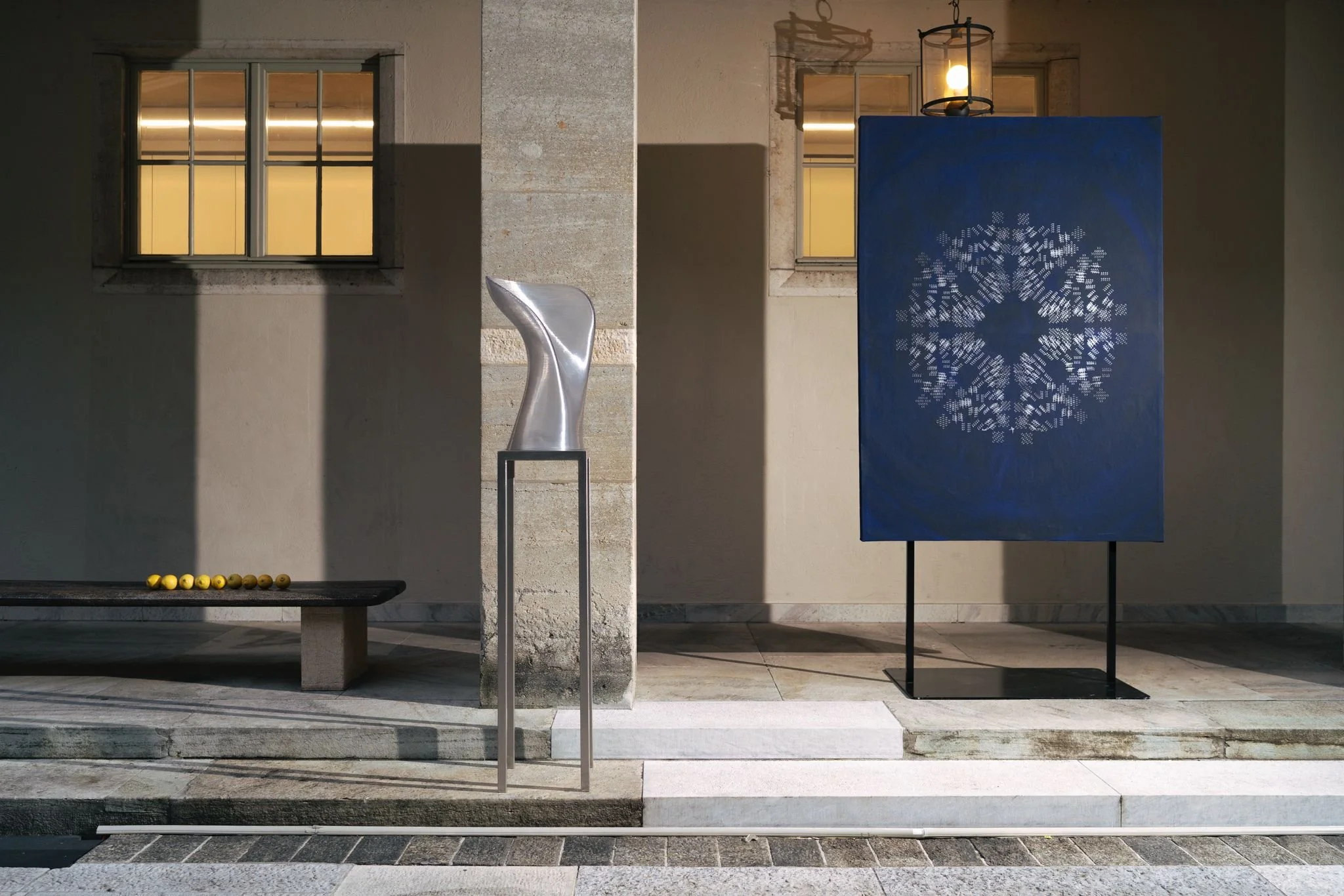 Exterior view of a modern building at night with two windows, a stone pillar, a bench with yellow balls, a metallic sculpture, a blue decorative panel with a geometric pattern, and a hanging lantern.