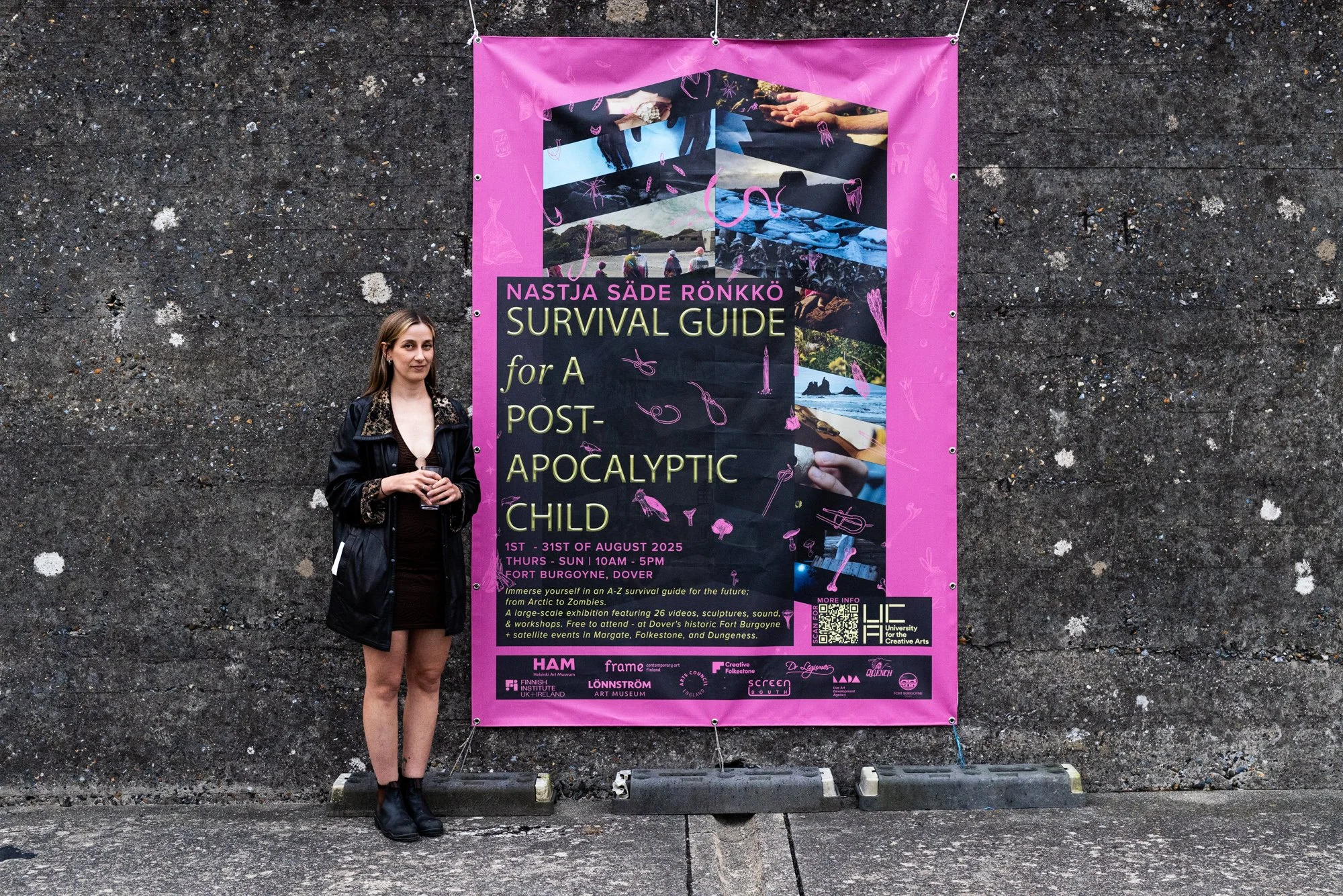 A woman stands next to a large pink poster on a dark, textured wall. The poster advertises an exhibition called 'Survival Guide for A Post-Apocalyptic Child,' scheduled from August 1st to August 31st, 2025, at Fort Burgoyne, Dover. The poster feature