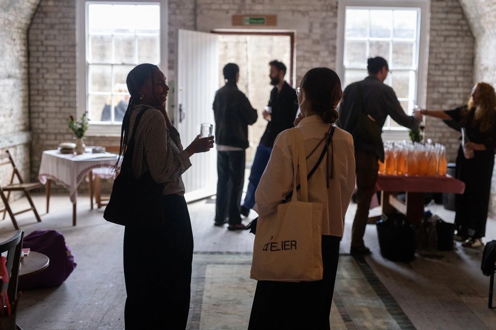 People socializing at an indoor gathering with brick walls and large windows, some holding drinks, and a table with bottles in the background.