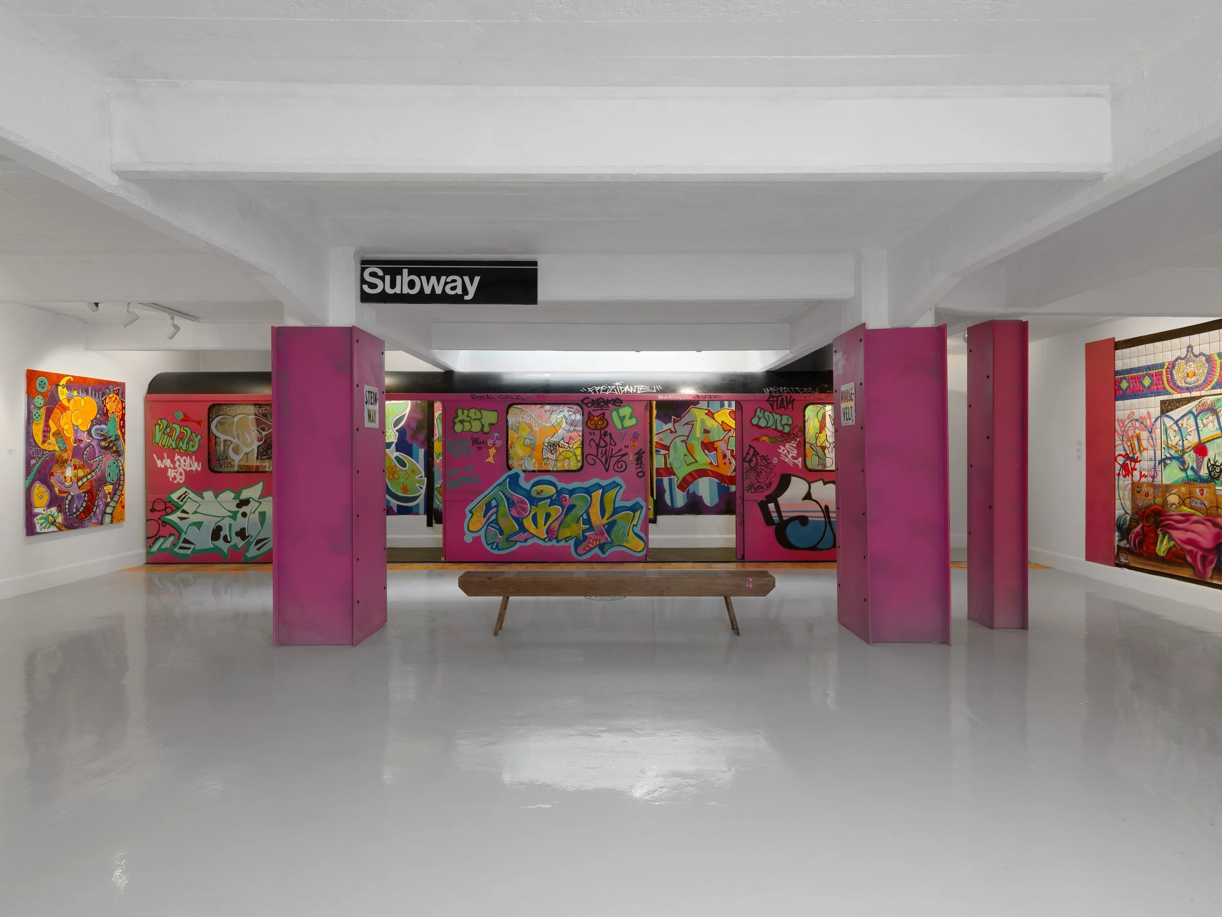 Art gallery featuring graffiti-style artwork displayed on pink panels and walls by Lady Pink, with a black and white 'Subway' sign hanging above in a white room.