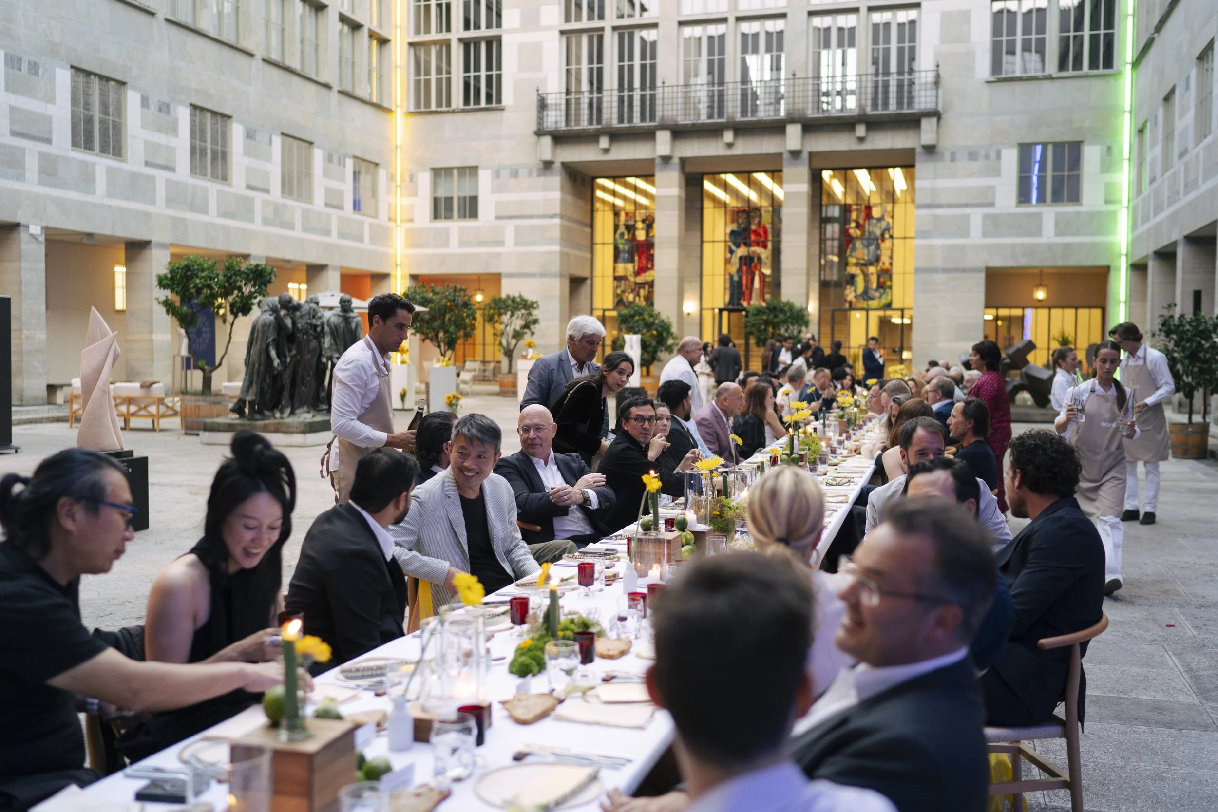 A large group of people seated at a long banquet table in a spacious, modern courtyard with glass windows, stained glass artwork, and greenery. The table is decorated with candles, flowers, and place settings.