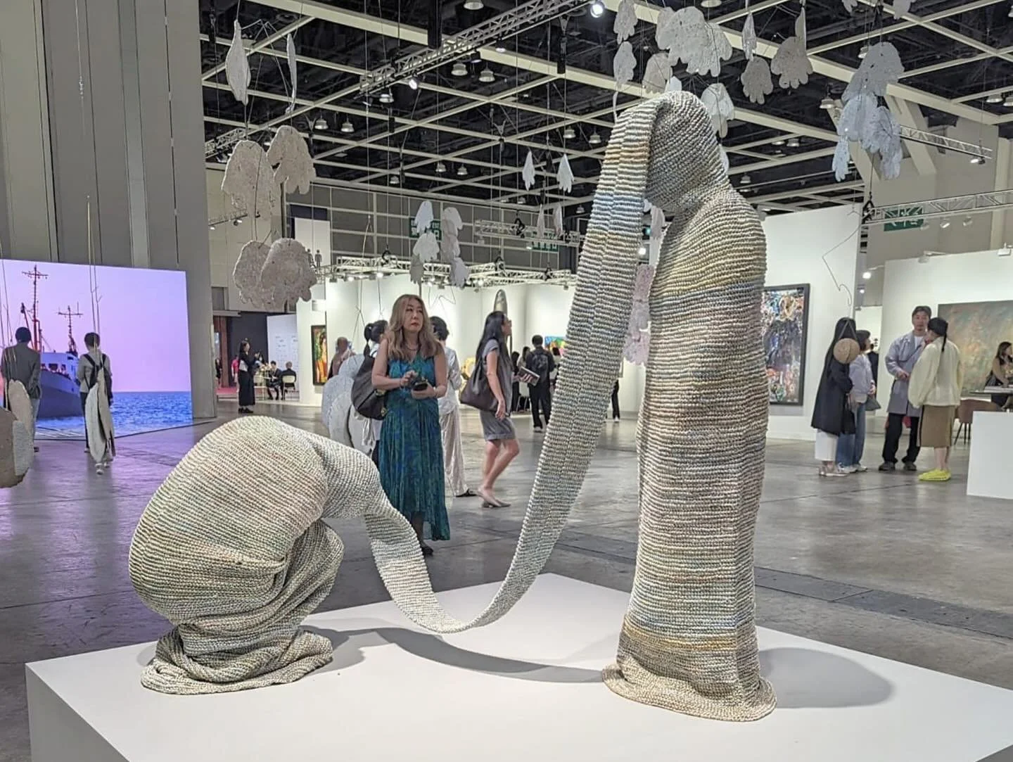 Art Basel Hong Kong, an art installation of a human figure with elongated limbs made from layered material, standing on a white platform at an art gallery, with visitors observing in the background.