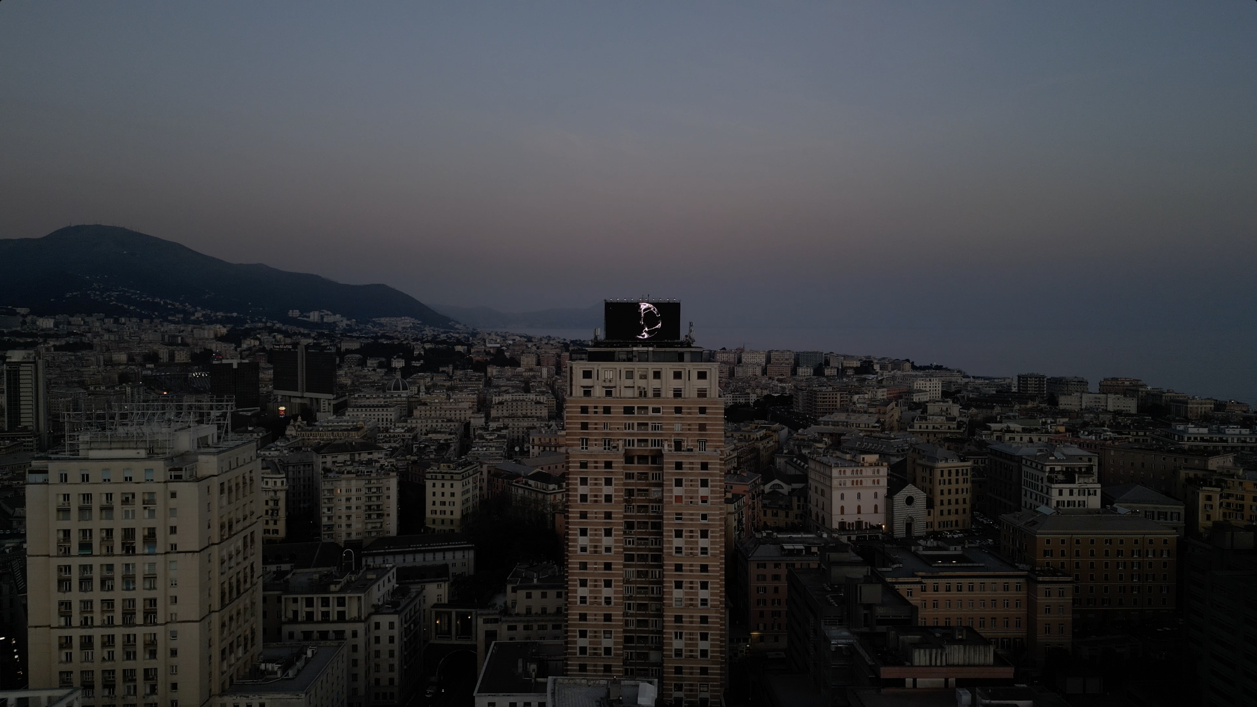 City skyline at dusk featuring numerous buildings, a large digital billboard on a building, mountains in the background, and a cloudy sky.