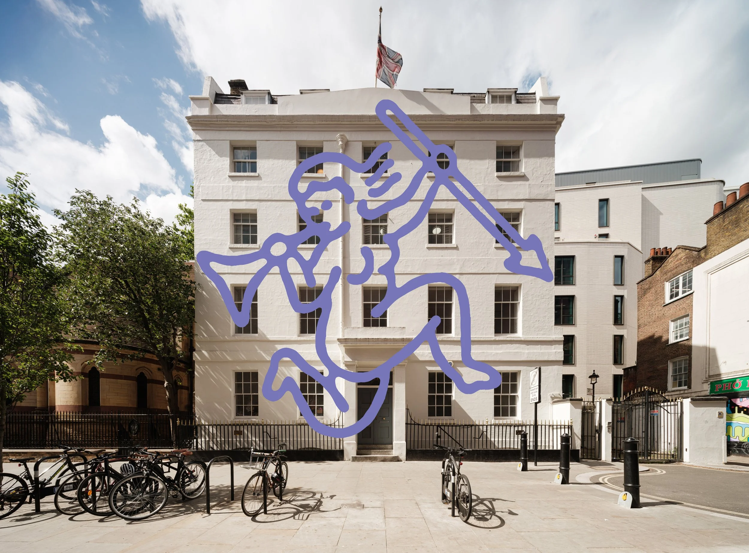 A white multi-story building with several windows, a flag on top, and a painted outline of a hand with a raised index finger in purple on its facade. In front, there is a small fence, bicycles, and trees on a sidewalk side under a partly cloudy sky.