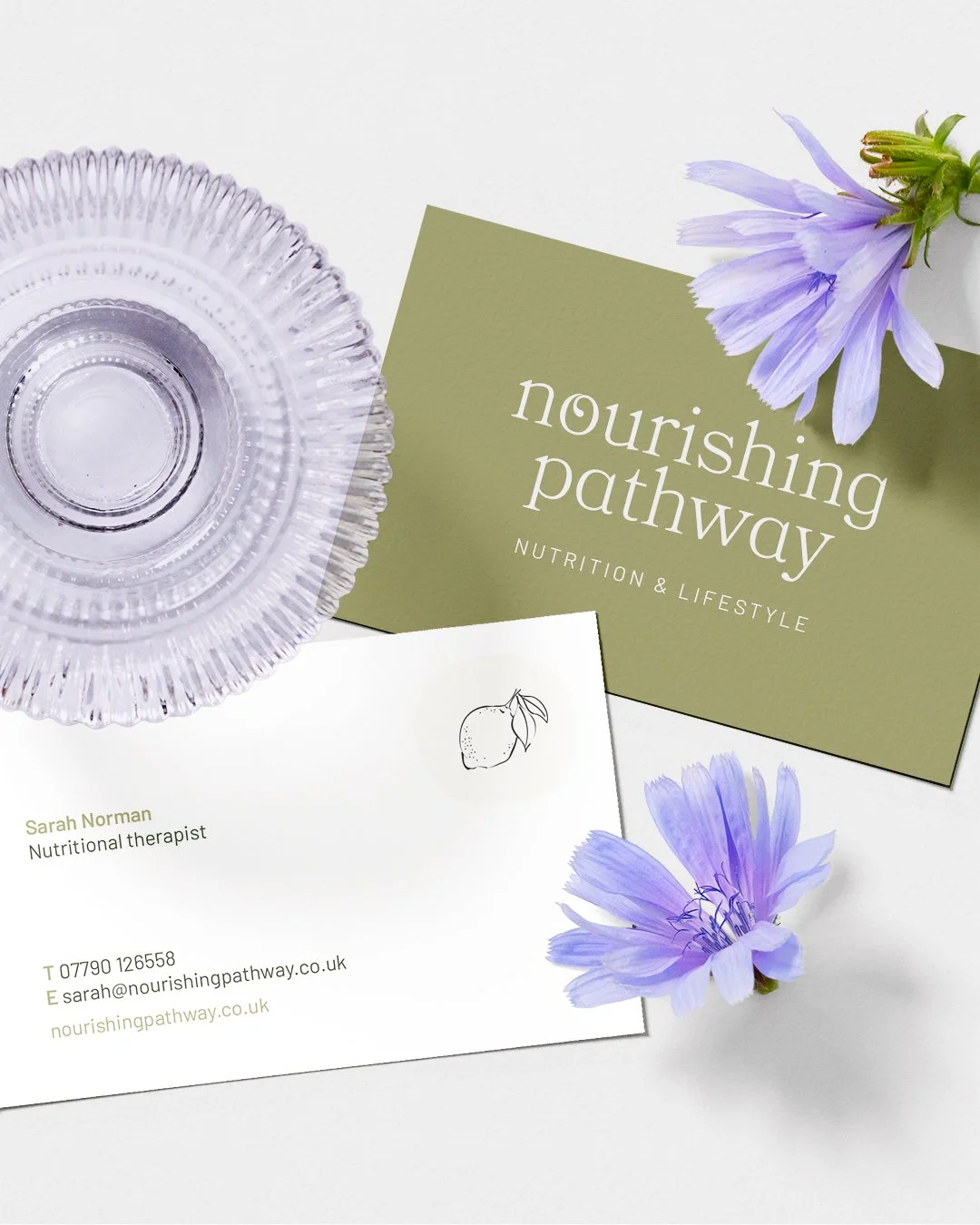 Business card for Sarah Norman, nutritional therapist, with a lemon illustration, placed next to a glass dish and purple flowers on a white background. The card includes contact details and website for Nourishing Pathway.