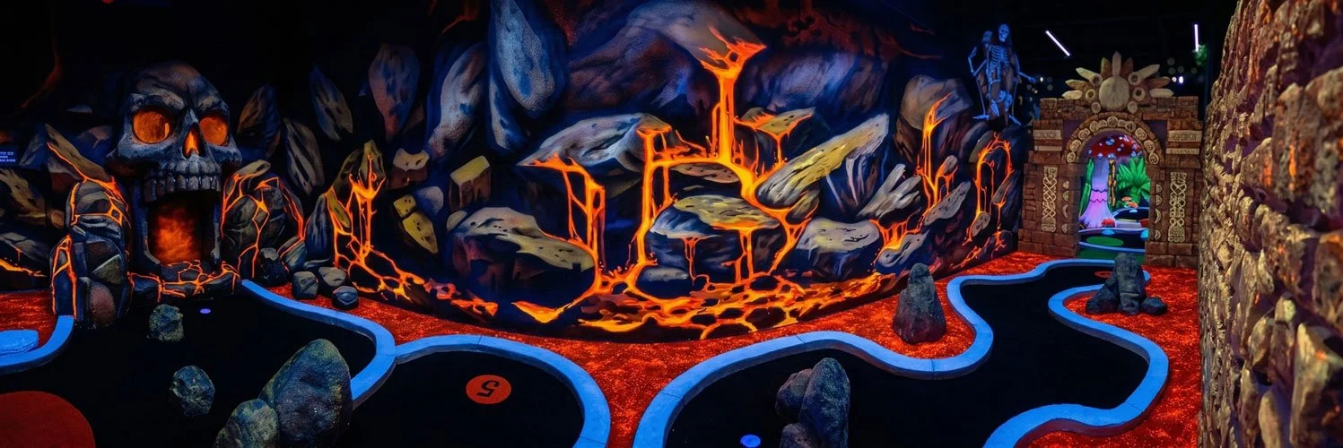 A vibrant, enchanting indoor blacklight miniature golf course designed by Charmed Playhouses. The fantastical setting features glowing 3D mushroom statues, a large gnarled tree, and elaborate stone structures. The walls are covered in striking, color