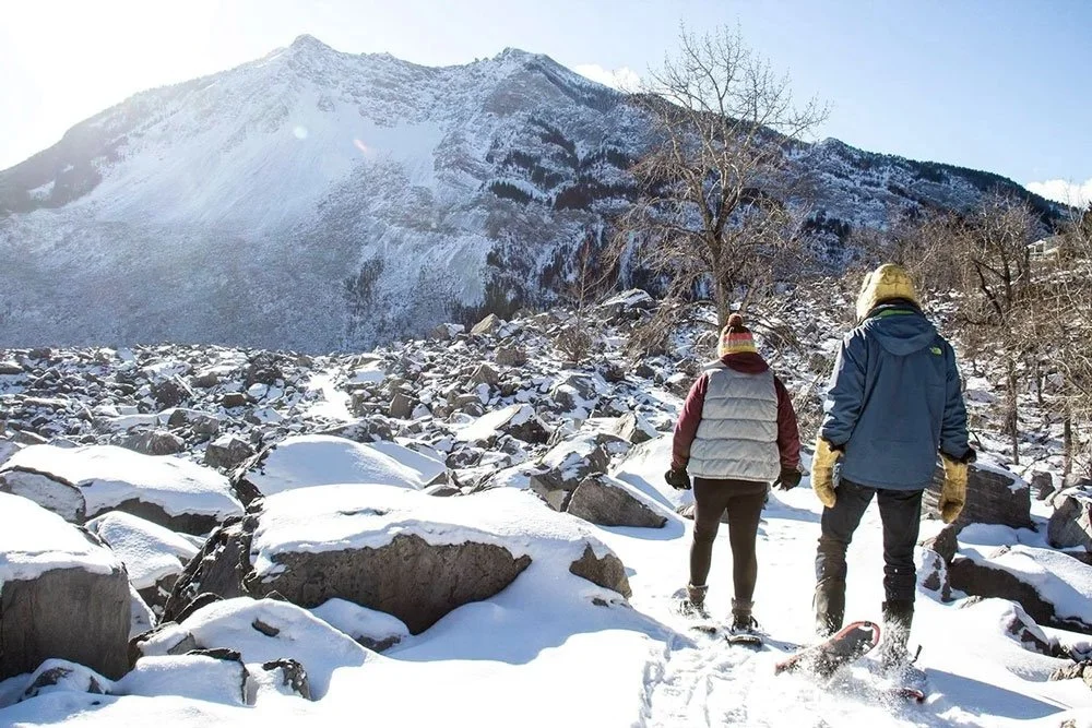 Two people hiking through a snowy, rocky landscape at the base of a snow-covered mountain during daytime.