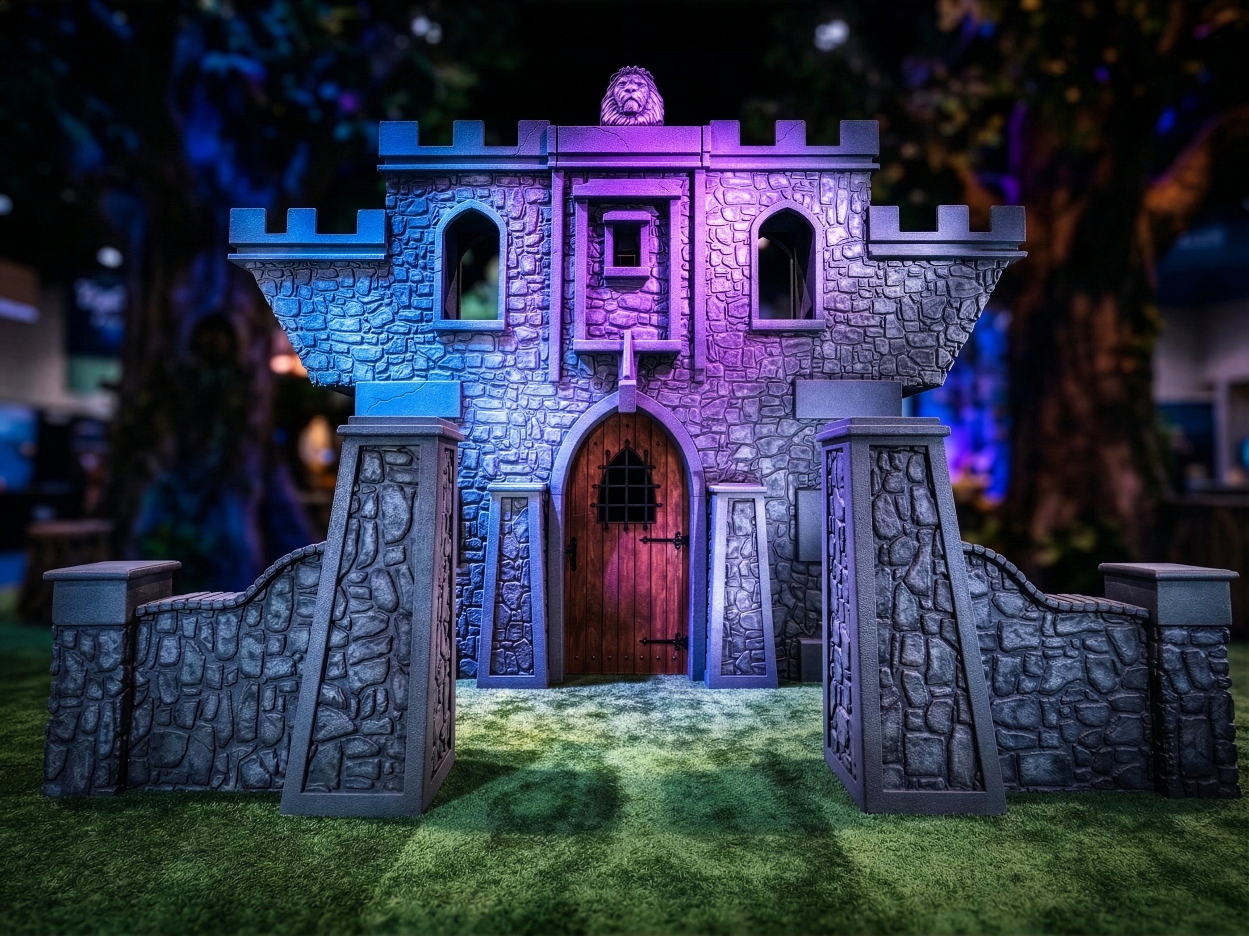 A brightly lit, purple and blue-lit grey stone castle structure with a large lion statue on top, a wooden arched doorway, and small crenelated turrets sits on a green, textured grassy ground, with blurred tree branches in the background by Charmed.