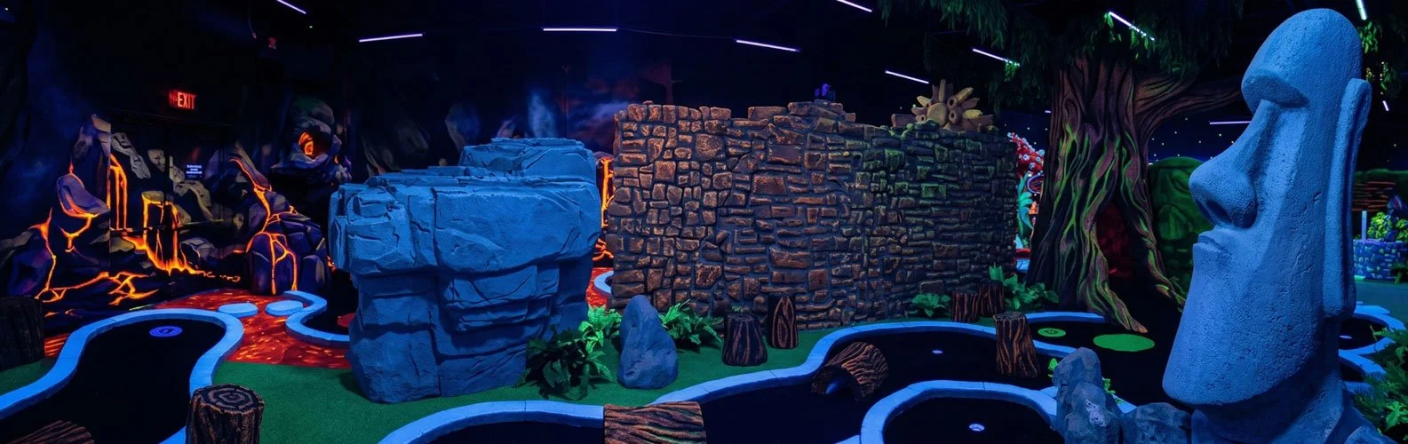 A vibrant, enchanting indoor blacklight miniature golf course designed by Charmed Playhouses. The fantastical setting features glowing 3D mushroom statues, a large gnarled tree, and elaborate stone structures. The walls are covered in striking, color