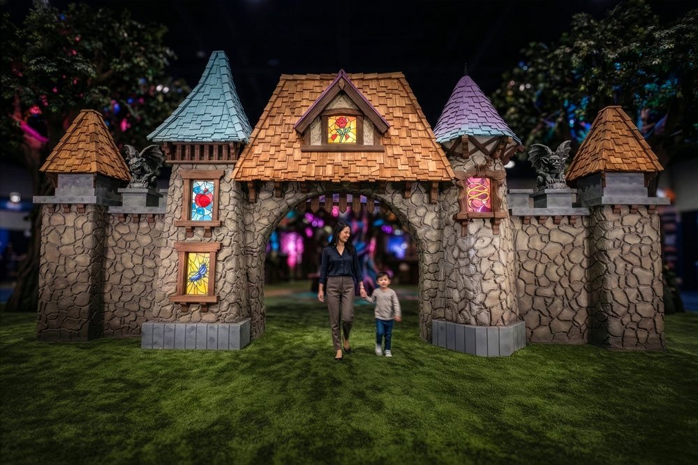 A brightly lit, purple and blue-lit grey stone castle structure with a large lion statue on top, a wooden arched doorway, and small crenelated turrets sits on a green, textured grassy ground, with blurred tree branches in the background by Charmed.