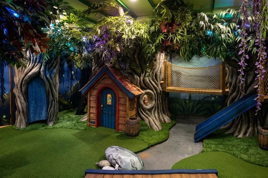 A custom indoor playroom by Charmed Playhouses in Lethbridge, Canada, recreates a magical woodland. It features large, twisted faux trees that form the structure, with a small, detailed wooden cottage nestled between them. The cottage has a blue door