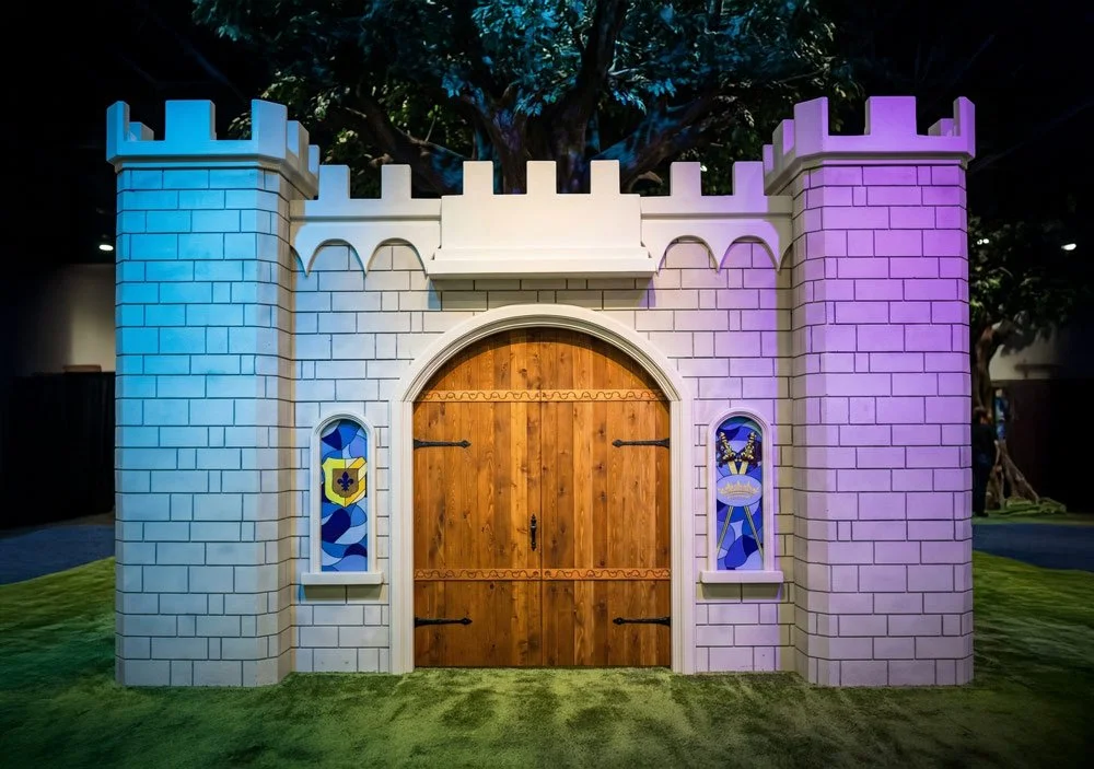 A brightly lit, purple and blue-lit grey stone castle structure with a large lion statue on top, a wooden arched doorway, and small crenelated turrets sits on a green, textured grassy ground, with blurred tree branches in the background by Charmed.