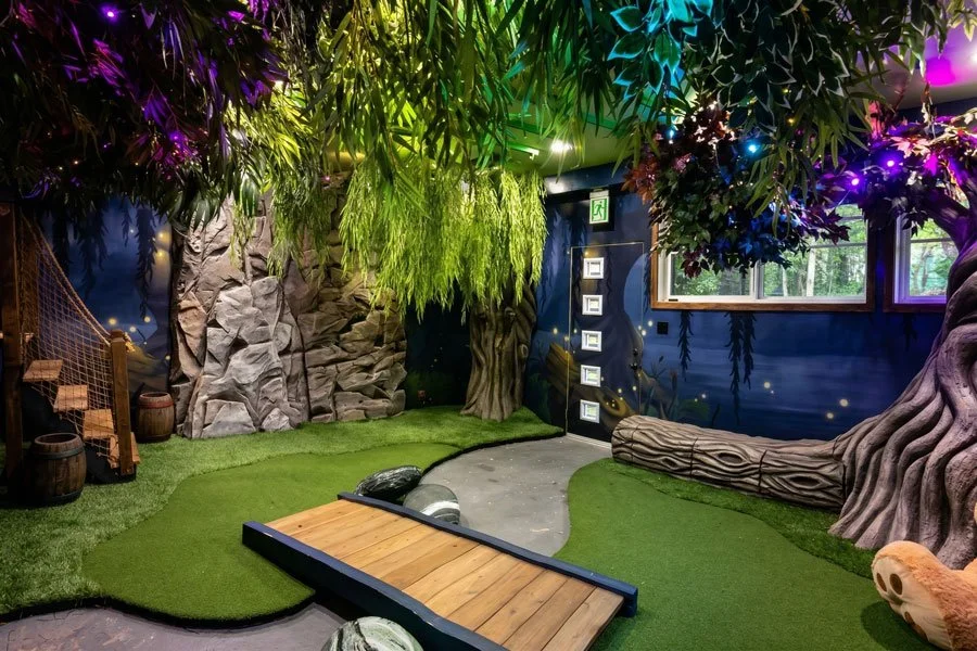 A custom indoor playroom by Charmed Playhouses in Lethbridge, Canada, recreates a magical woodland. It features large, twisted faux trees that form the structure, with a small, detailed wooden cottage nestled between them. The cottage has a blue door
