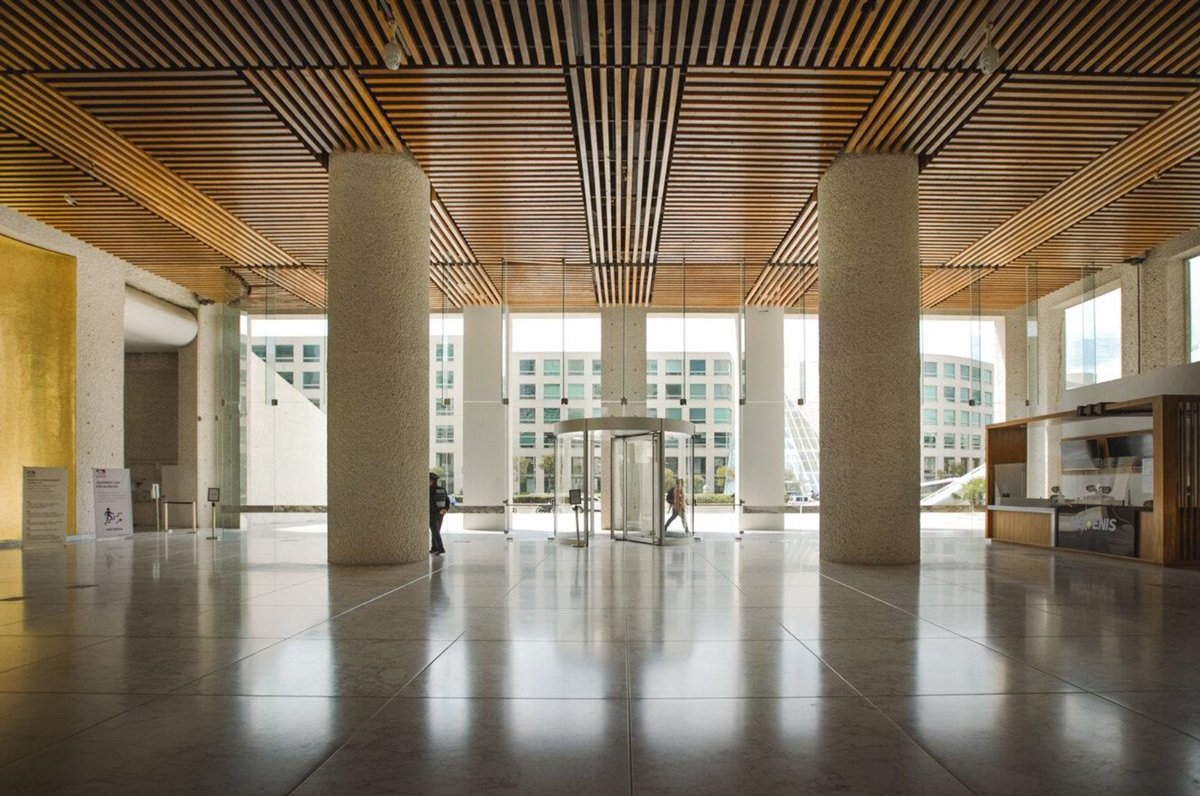 El lobby de Torre Arcos Bosques: columnas de concreto, plafón de madera y transparencia hacia la plaza.