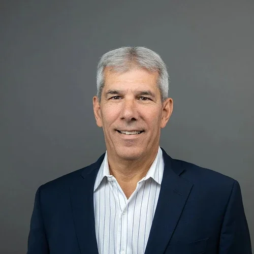 Headshot of a middle-aged man with gray hair, wearing a dark suit jacket and a light striped dress shirt, smiling against a plain gray background.