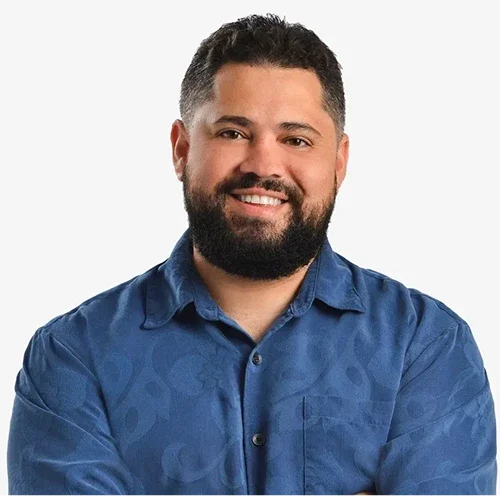 A smiling man with dark hair and a beard, wearing a blue button-up shirt, against a white background.