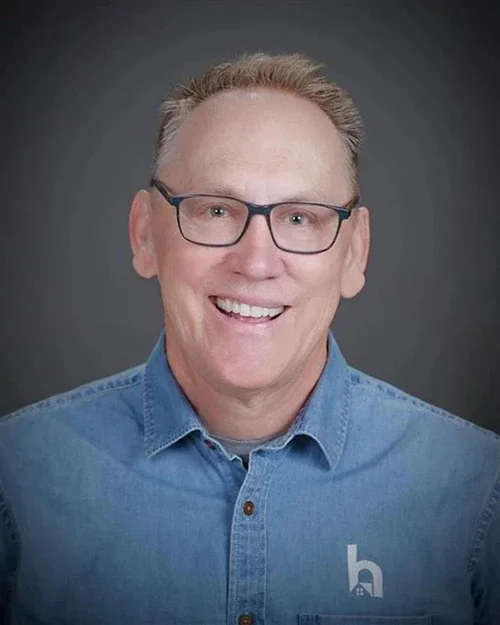 Headshot of a middle-aged man with light skin, glasses, short light brown hair, smiling, wearing a blue denim shirt with a small logo on the chest, dark gray background.