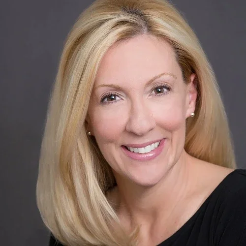 A smiling blonde woman with straight hair wearing a black top and pearl earrings.