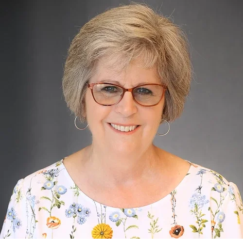 A middle-aged woman with short, blonde hair, wearing glasses and hoop earrings, smiling at the camera, wearing a white floral top, in front of a plain gray background.