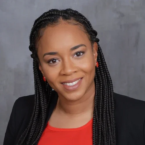 Close-up of a smiling woman with braided hair, wearing a black blazer and a red top, against a gray background.