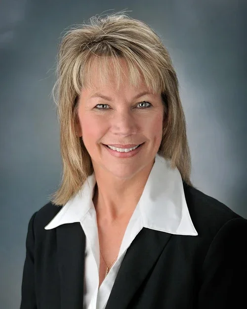 A professional headshot of a smiling middle-aged woman with short blonde hair, blue eyes, wearing a black blazer and white collared shirt against a gray background.