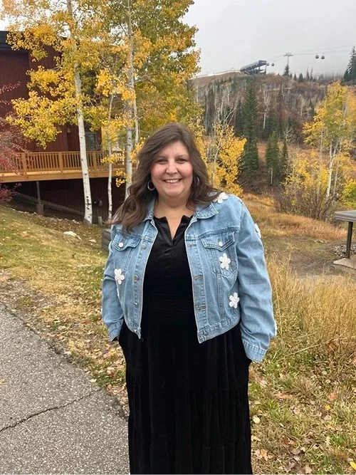 Woman in black dress and denim jacket standing outdoors with fall foliage and ski lift in the background.