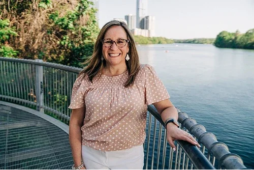 A woman with shoulder-length brown hair and glasses smiling on a bridge over a river, with city buildings and green trees in the background.