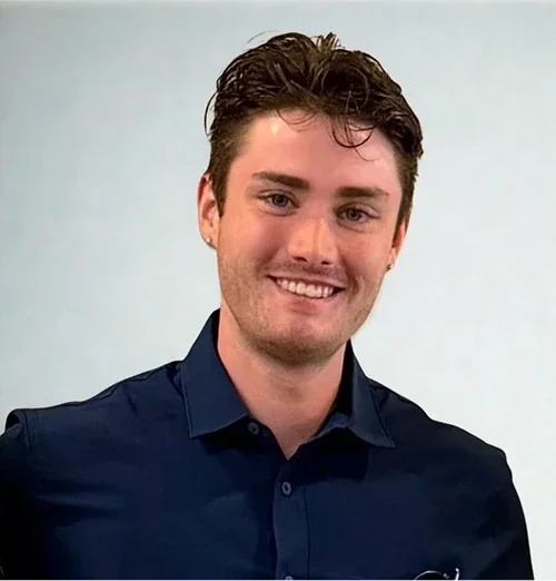 A young man with dark hair, blue eyes, and light skin, smiling and wearing a dark blue collared shirt against a plain light background.