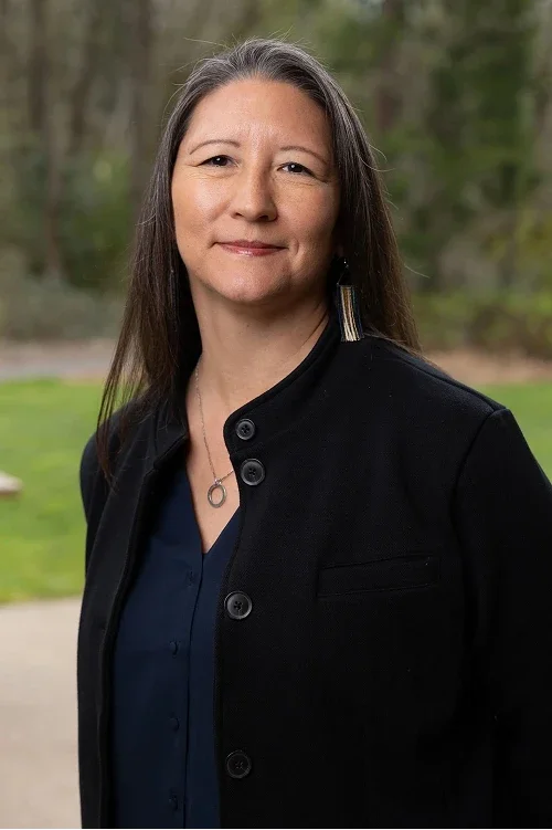 A woman with long dark hair and earrings standing outdoors in front of trees and grass, wearing a dark blazer over a navy shirt, smiling subtly.