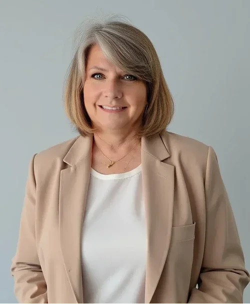 A middle-aged woman with shoulder-length light brown hair, smiling, wearing a beige blazer over a white top against a plain light gray background.